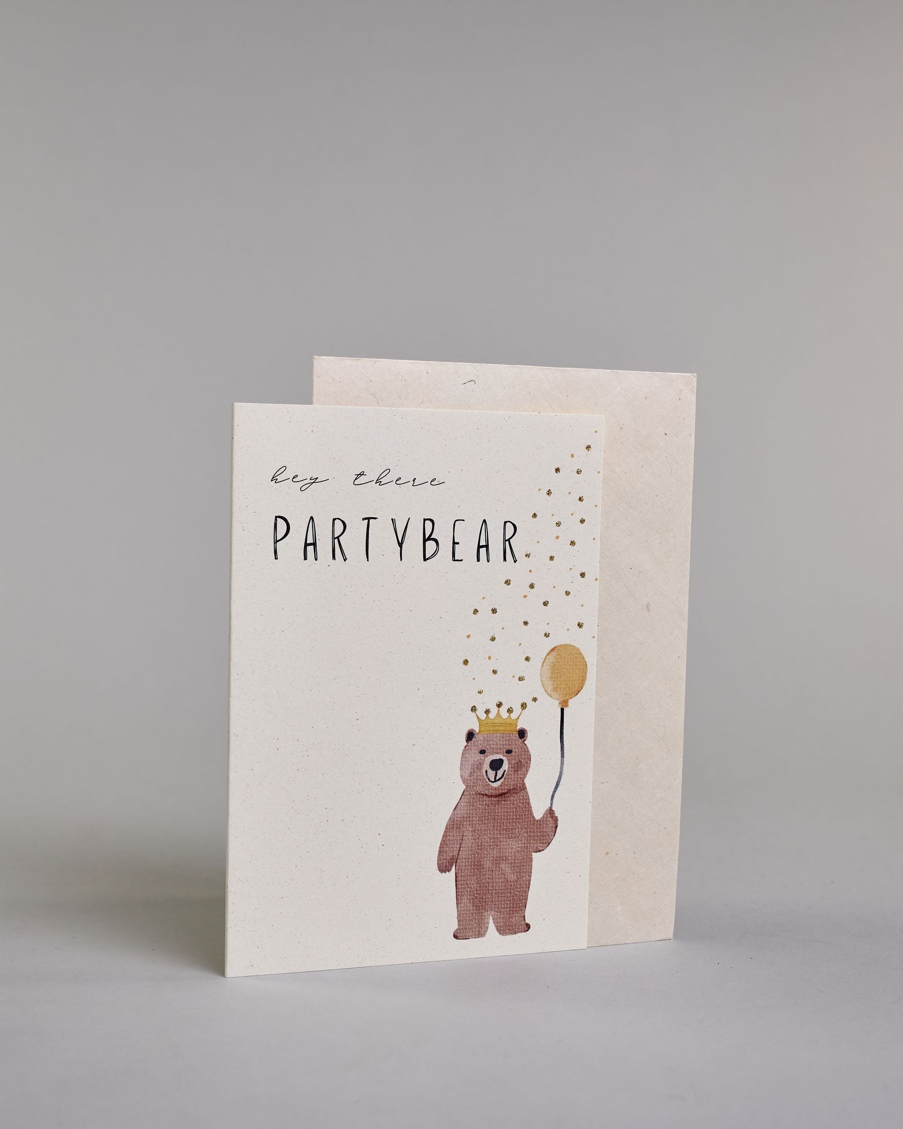 Glückwunschkarte auf Englisch zum Geburtstag mit schwarzer Kalligrafieaufschrift "hey there Partybear", mit einem gezeichneten braunen Bären der eine Krone aufhat und einen gelben Luftbalon in der rechten Pfote hält auf hellem Hintergrund. Oben steht in dünner geschwungener Schrift "hey there", darunter "PARTYBEAR" in gerader dünner Kalligrafieschrift, rechts unten steht der Bäre, hinter ihm steigen Konfettiähnlich gelbe und goldglitzernde Punkte bis zur rechten oben Ecke der Karte auf. Dazu ein grün-golde…