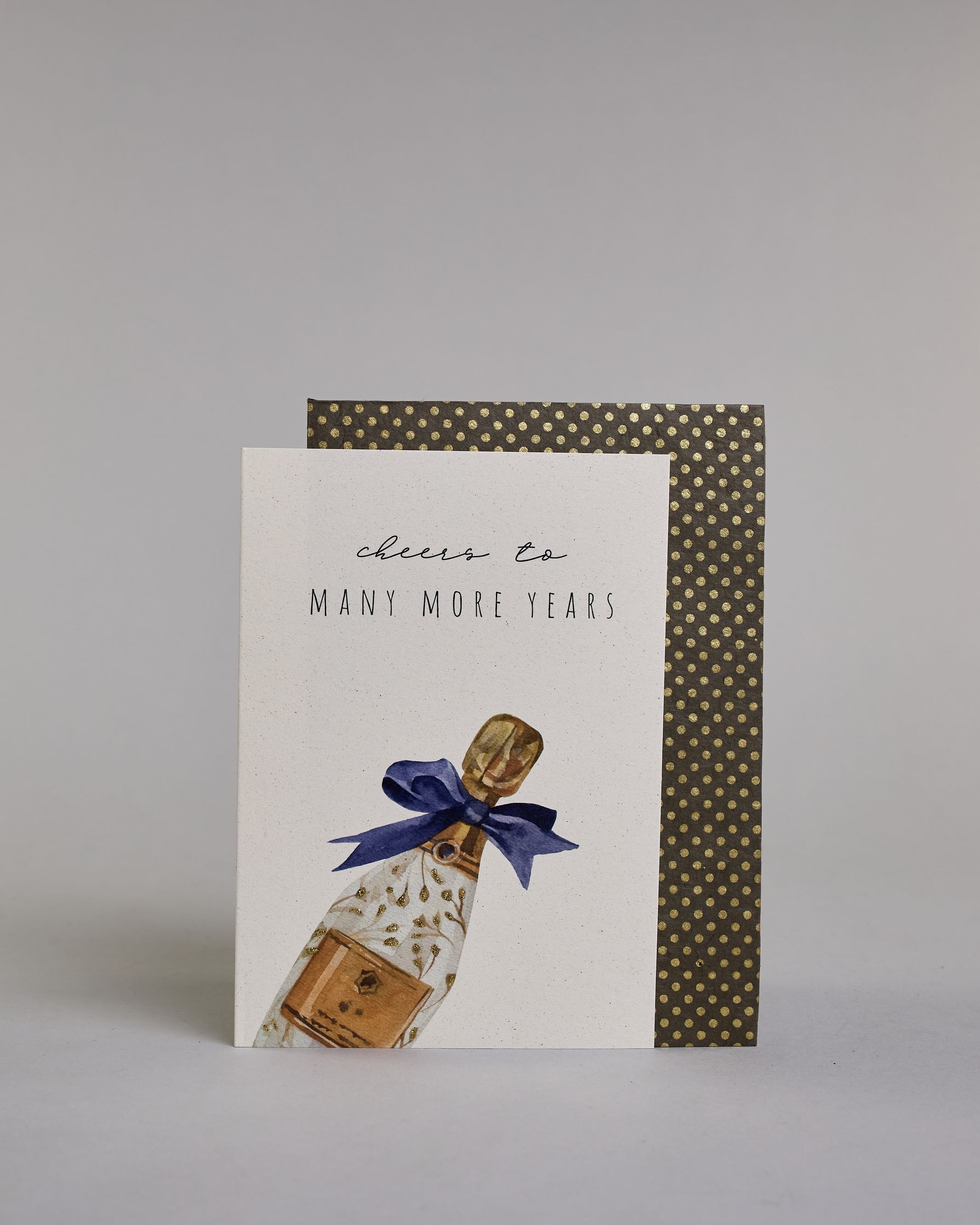 Glückwunschkarte auf Englisch zum Geburtstag mit schwarzer Kalligrafieaufschrift "cheers to many more years", mit einer gezeichneten Champagnerflasche mit goldenen Blättern als verzierung, goldenem Ettikett, goldener Flaschenhals- und Kopf und deiner blauen Masche um den Flaschenhals auf hellem Hintergrund. Oben steht in dünner geschwungener Schrift "cheers to", darunter "MANY MORE YEARS" in gerader dünner Kalligrafieschrift. Darunter kommt die falsche nach rechts geneigt von der linken Kante der Karte her…