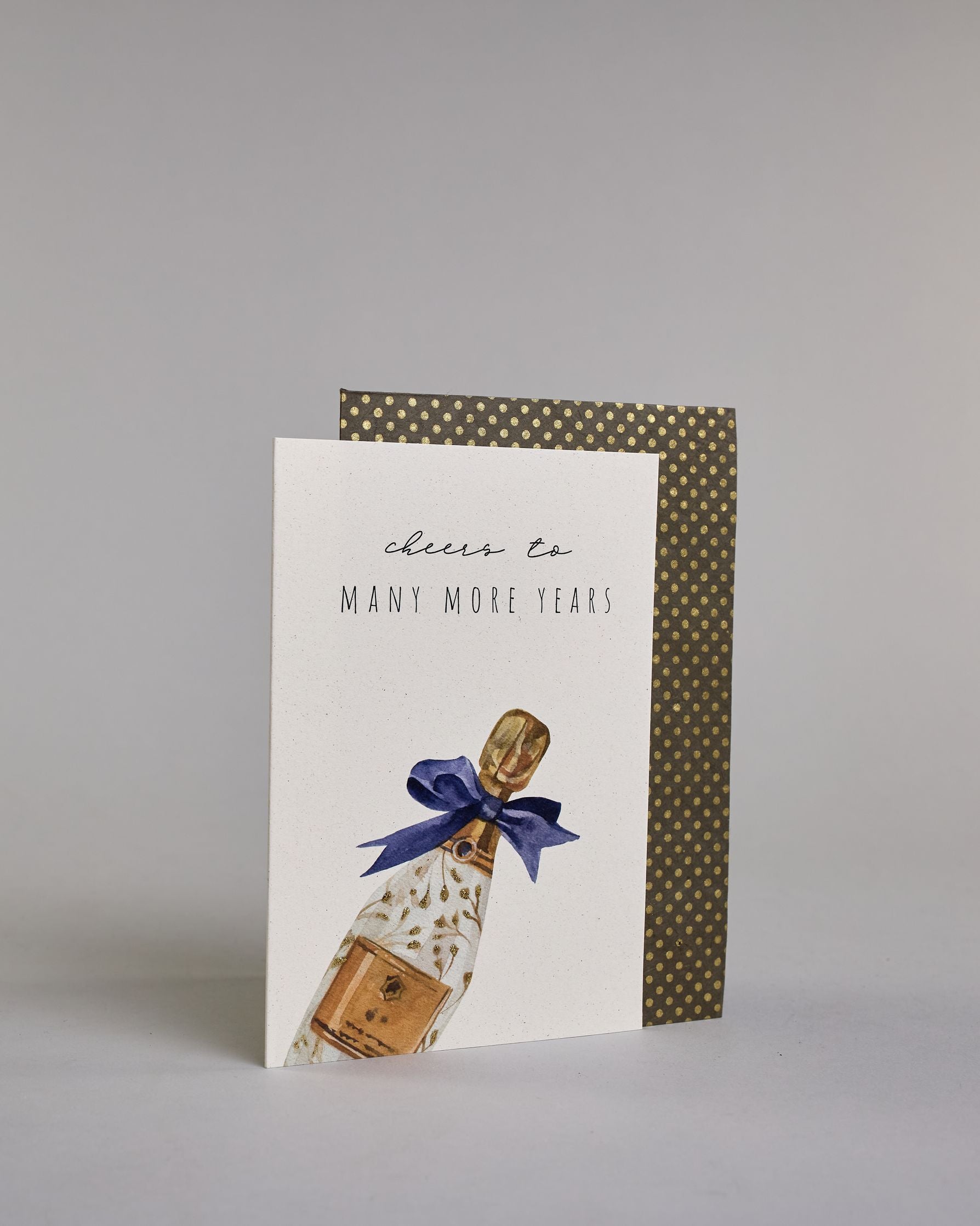 Glückwunschkarte auf Englisch zum Geburtstag mit schwarzer Kalligrafieaufschrift "cheers to many more years", mit einer gezeichneten Champagnerflasche mit goldenen Blättern als verzierung, goldenem Ettikett, goldener Flaschenhals- und Kopf und deiner blauen Masche um den Flaschenhals auf hellem Hintergrund. Oben steht in dünner geschwungener Schrift "cheers to", darunter "MANY MORE YEARS" in gerader dünner Kalligrafieschrift. Darunter kommt die falsche nach rechts geneigt von der linken Kante der Karte her…