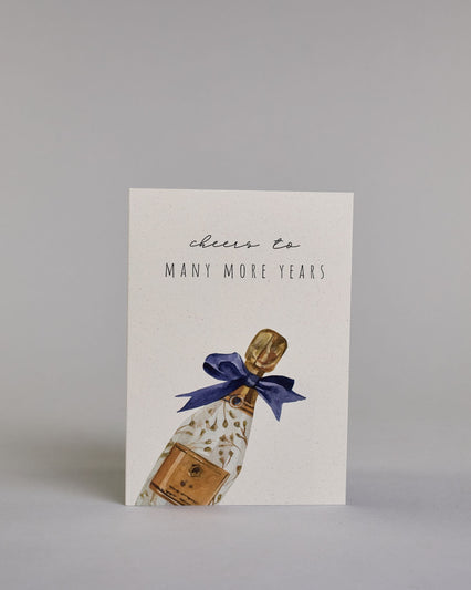 Glückwunschkarte auf Englisch zum Geburtstag mit schwarzer Kalligrafieaufschrift "cheers to many more years", mit einer gezeichneten Champagnerflasche mit goldenen Blättern als verzierung, goldenem Ettikett, goldener Flaschenhals- und Kopf und deiner blauen Masche um den Flaschenhals auf hellem Hintergrund. Oben steht in dünner geschwungener Schrift "cheers to", darunter "MANY MORE YEARS" in gerader dünner Kalligrafieschrift. Darunter kommt die falsche nach rechts geneigt von der linken Kante der Karte her…