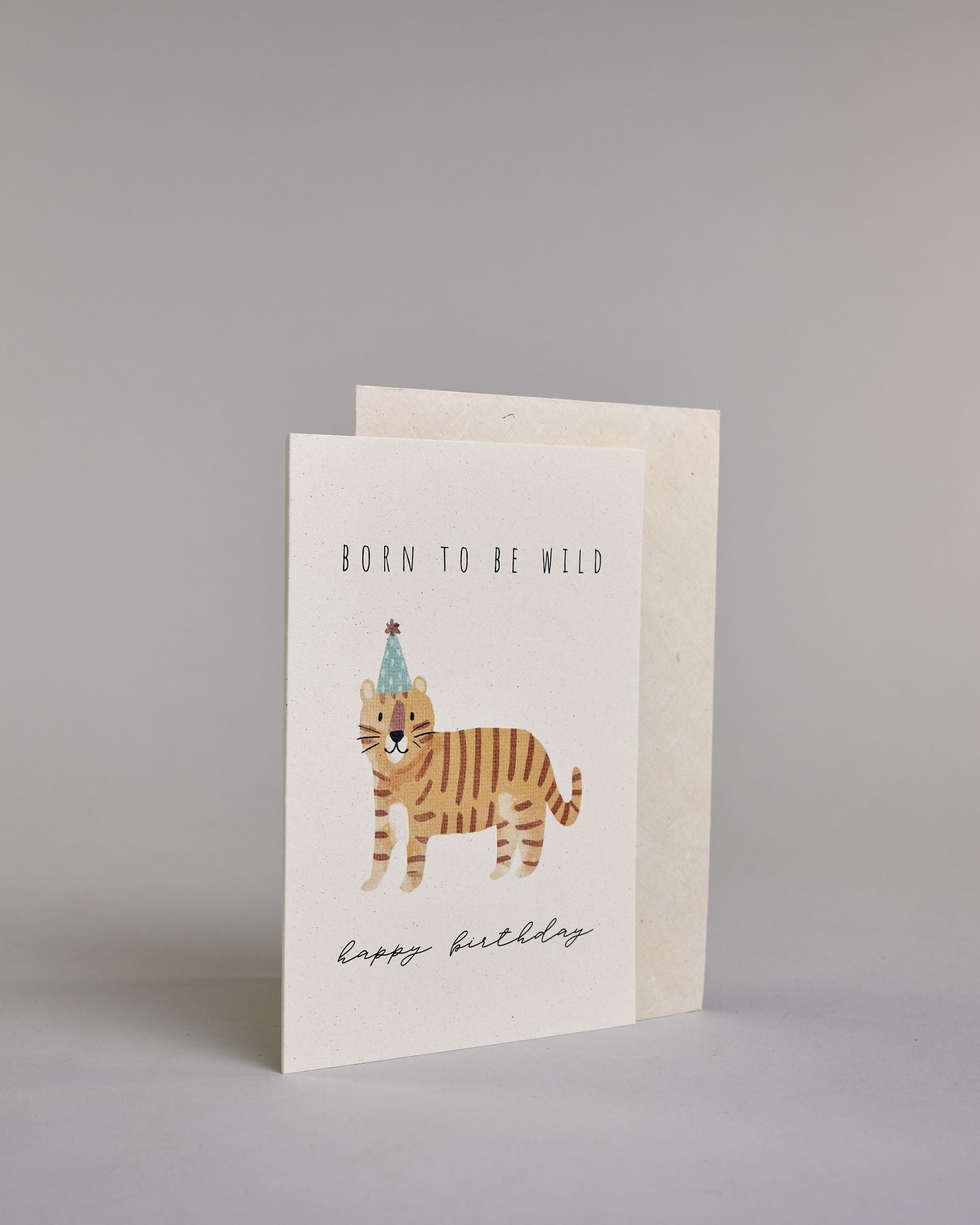 Glückwunschkarte auf Englisch zum Geburtstag mit schwarzer Kalligrafieaufschrift "born to be wild happy birthday", mit einer kindlich gezeichneten orange getigerten Katze mit blauem Partyhut auf hellem Hintergrund. Oben steht in dünner gerader Schrift "BORN TO BE WILD", darunter ist mittig die Katze, darunter "happy birthday" in geschwungener dünner Kalligrafieschrift. Dazu ein weißes handgeschöpftes Kuvert. #3