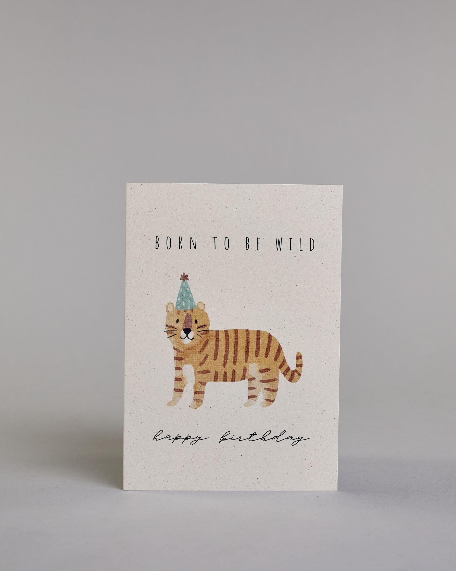 Glückwunschkarte auf Englisch zum Geburtstag mit schwarzer Kalligrafieaufschrift "born to be wild happy birthday", mit einer kindlich gezeichneten orange getigerten Katze mit blauem Partyhut auf hellem Hintergrund. Oben steht in dünner gerader Schrift "BORN TO BE WILD", darunter ist mittig die Katze, darunter "happy birthday" in geschwungener dünner Kalligrafieschrift. Dazu ein weißes handgeschöpftes Kuvert. #1