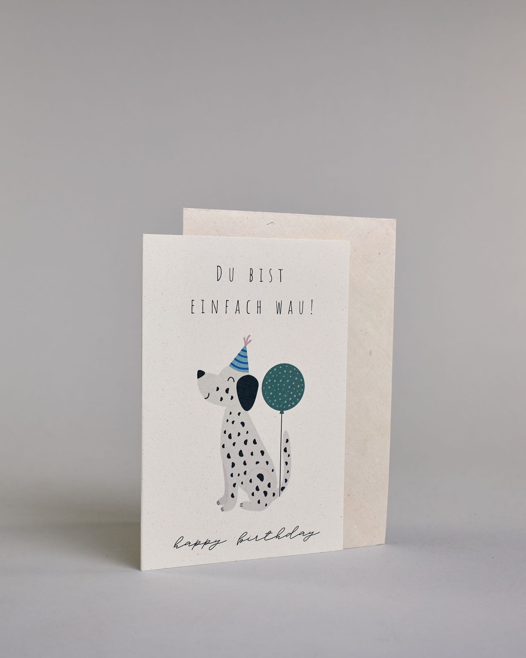 Glückwunschkarte zum Geburtstag mit schwarzer Kalligrafieaufschrift "Du bist einfach Wau! Happy Birthday", mit einem kindlich gezeichneten Dalmatiner mit blauem Partyhut an dessen Schwanz ein petrolfarbener Balon festgebunden ist auf hellem Hintergrund. Oben steht in dünner gerader Schrift "DU BIST", darunter "EINFACH WAU!", darunter ist mittig der schwarzweiß gepunktete Hund, darunter "happy birthday" in geschwungener dünner Kalligrafieschrift. Dazu ein weißes handgeschöpftes Kuvert. #3