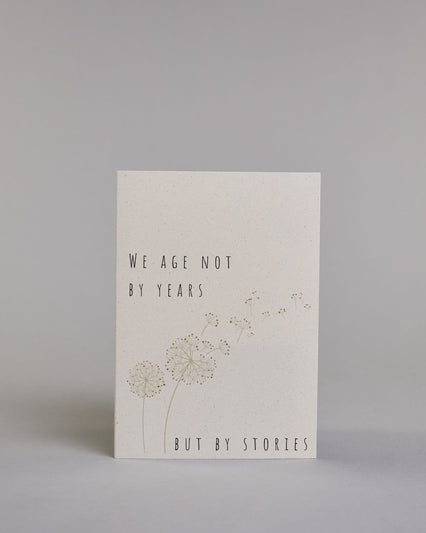 Glückwunschkarte auf Englisch zum Geburtstag mit schwarzer Kalligrafieaufschrift "We age not by years but by stories", mit zwei gezeichneten Pusteblumen deren Samen verweht werden auf hellem Hintergrund. Oben links steht in dünner gerader Schrift "WE AGE NOT", darunter "BY YEARS", darunter von links nach rechtswandernd die goldverzierte Pusteblume, darunter rechts "BUT BY STORIES" in gerader dünner Kalligrafieschrift. Dazu ein weißes handgeschöpftes Kuvert. #1