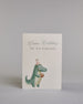 Glückwunschkarte auf Englisch zum Geburtstag mit blauer und schwarzer Kalligrafieaufschrift "Happy Birthday you old Dinosaur", mit einem kindlich genzeichnetem grünen Dinosaurier mit pinkem Partyhut und einem orangen Geschenk in den Händen auf hellem Hintergrund. Oben steht in zwei Zeilen "Happy Birthday" in dünner geschwungener blauer Schreibschrift, darunter "YOU OLD DINOSAUR" in dünner gerader schwarzer Kalligrafieschrift, darunter ist auf dem linken Drittel der Karte der Dinosaurier. Dazu ein blau-weiß…