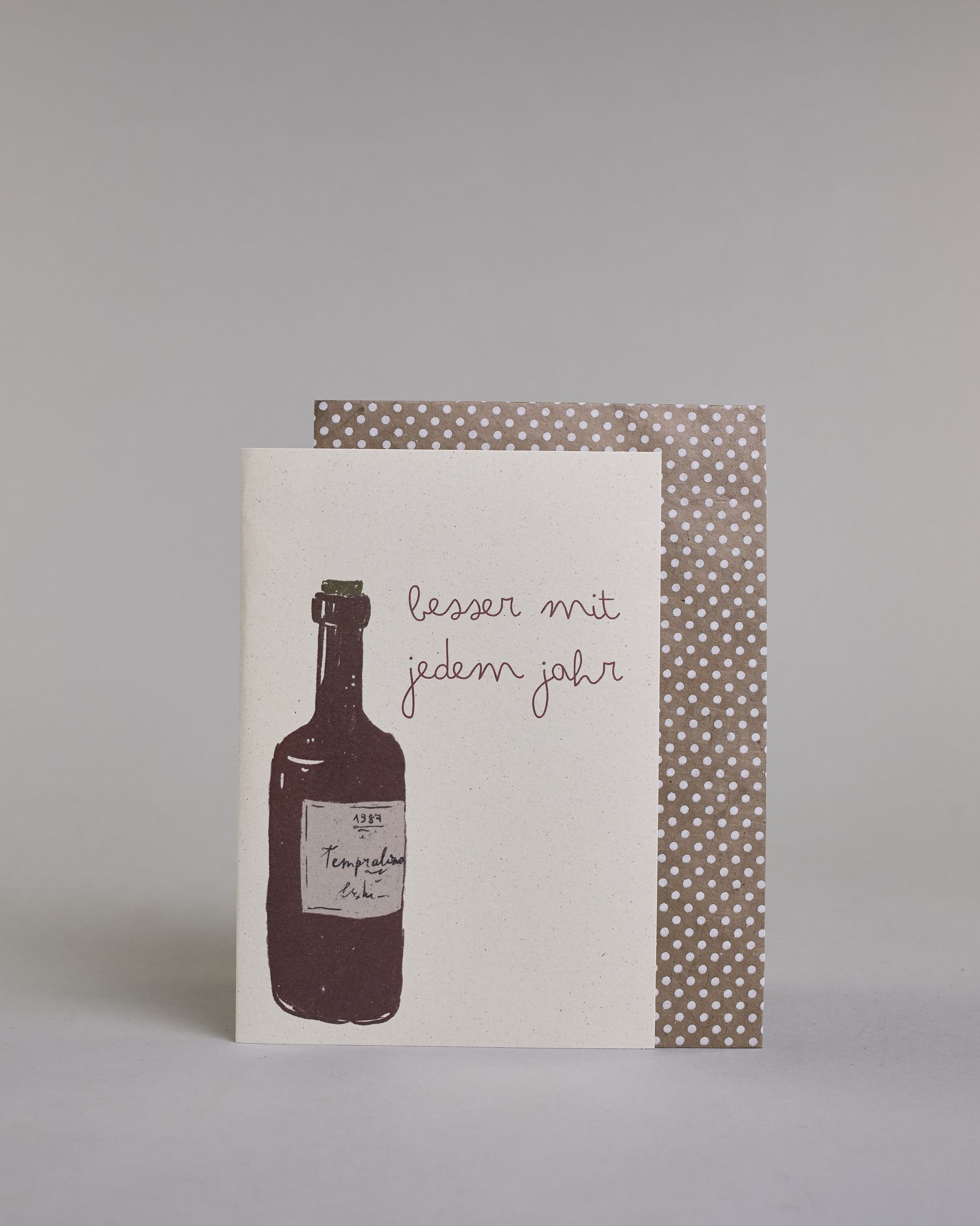 Glückwunschkarte zum Geburtstag mit brauner Kalligrafieaufschrift "besser mit jedem Jahr", mit einer genzeichneten braunen Weinflasche mit beigen Ettikett auf hellem Hintergrund. Oben steht in zwei Zeilen "besser mit", darunter "jedem jahr" beides in dünner geschwungener Schreibschrift, links davon die Weinflasche, die Schrift befindet sich neben dem Flaschenhals. Dazu ein beige-weißes handgeschöpftes Kuvert. #2