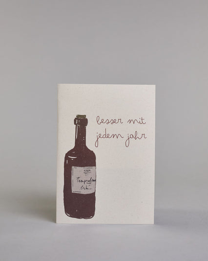 Glückwunschkarte zum Geburtstag mit brauner Kalligrafieaufschrift "besser mit jedem Jahr", mit einer genzeichneten braunen Weinflasche mit beigen Ettikett auf hellem Hintergrund. Oben steht in zwei Zeilen "besser mit", darunter "jedem jahr" beides in dünner geschwungener Schreibschrift, links davon die Weinflasche, die Schrift befindet sich neben dem Flaschenhals. Dazu ein beige-weißes handgeschöpftes Kuvert. #1