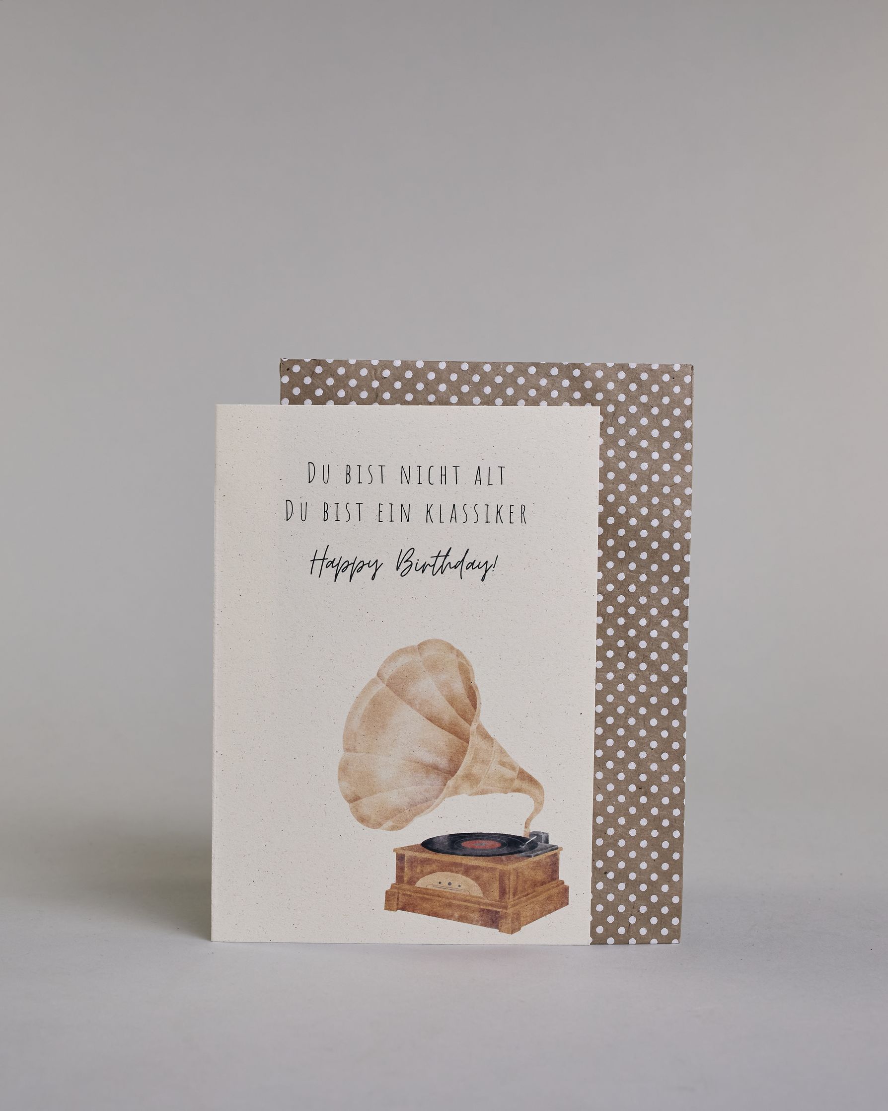 Glückwunschkarte zum Geburtstag mit schwarzer Kalligrafieaufschrift "Du bist nicht alt du bist ein Klassiker Happy Birthday!", mit einem genzeichneten klassischen braunen PLattenspieler mit einer schwarzen Platte auf hellem Hintergund. Oben steht in drei Zeilen "DU BIST NICHT ALT", darunter "DU BIST EIN KLASSIKER" beides in dünner gerader Kalligrafieschrift, darunter "Happy Birthday!" in geschwungener dünner Handschrift und darunter im rechten Drittel der Karte ist der Plattenspieler. Dazu ein beige-weißes…