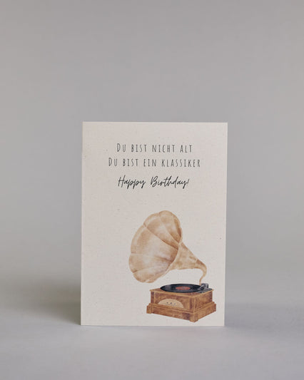 Glückwunschkarte zum Geburtstag mit schwarzer Kalligrafieaufschrift "Du bist nicht alt du bist ein Klassiker Happy Birthday!", mit einem genzeichneten klassischen braunen PLattenspieler mit einer schwarzen Platte auf hellem Hintergund. Oben steht in drei Zeilen "DU BIST NICHT ALT", darunter "DU BIST EIN KLASSIKER" beides in dünner gerader Kalligrafieschrift, darunter "Happy Birthday!" in geschwungener dünner Handschrift und darunter im rechten Drittel der Karte ist der Plattenspieler. Dazu ein beige-weißes…