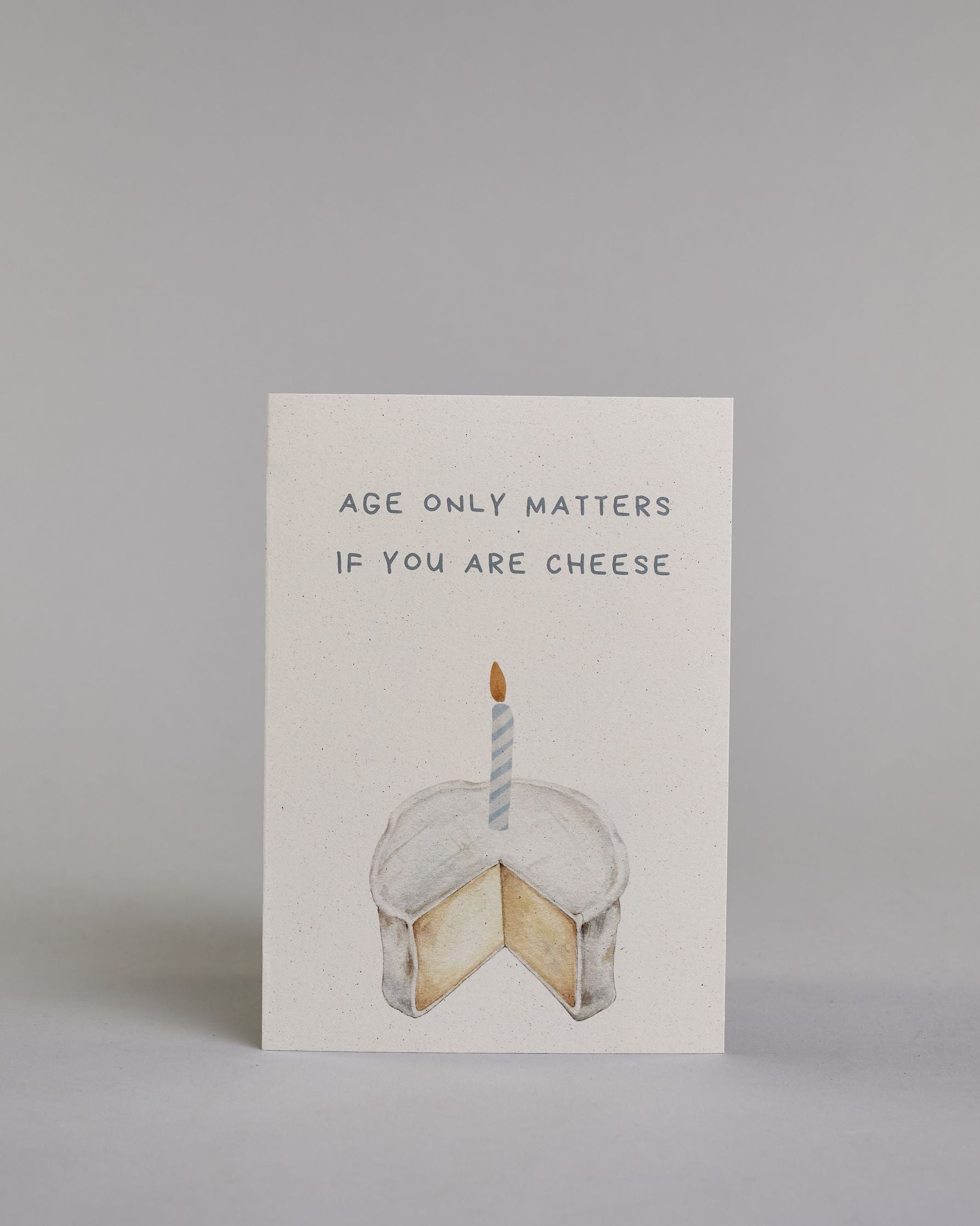 Glückwunschkarte auf Englisch zum Geburtstag mit blauer Kalligrafieaufschrift "age only matters if you are cheese", mit einem genzeichnetem Camembert in dem eine blau gestreifte Kerze steckt auf hellem Hintergund. Oben steht in zwei Zeilen "AGE ONLY MATTERS", darunter "IF YOU ARE CHEESE" beides in Blockbuchstaben und darunter mittig der Käse. Dazu ein blau-weißes handgeschöpftes Kuvert. #1