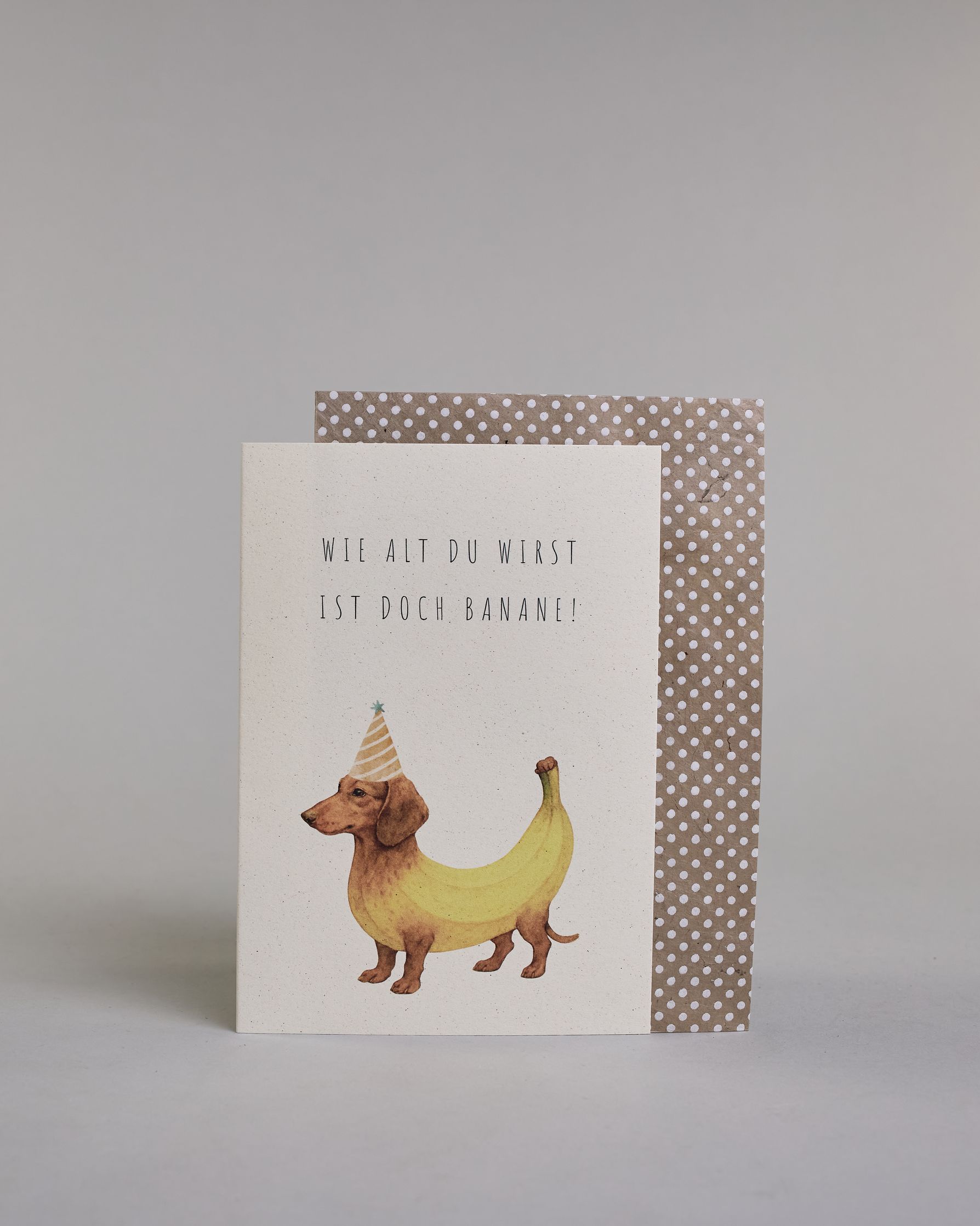 Glückwunschkarte zum Geburtstag mit schwarzer Kalligrafieaufschrift "Wie alt du wirst ist doch Banane!", mit einem genzeichnetem brauner Dackel dessen Körper aussieht wie eine gelbe Banane und einem gelb-orangen Partyhut auf dem Kopf auf hellem Hintergund. Oben steht in zwei Zeilen "WIE ALT DU WIRST", darunter "IST DOCH BANANE!" beides in dünner gerader Kalligrafieschrift und darunter mittig der Bananenhund. Dazu ein beige-weißes handgeschöpftes Kuvert. #2