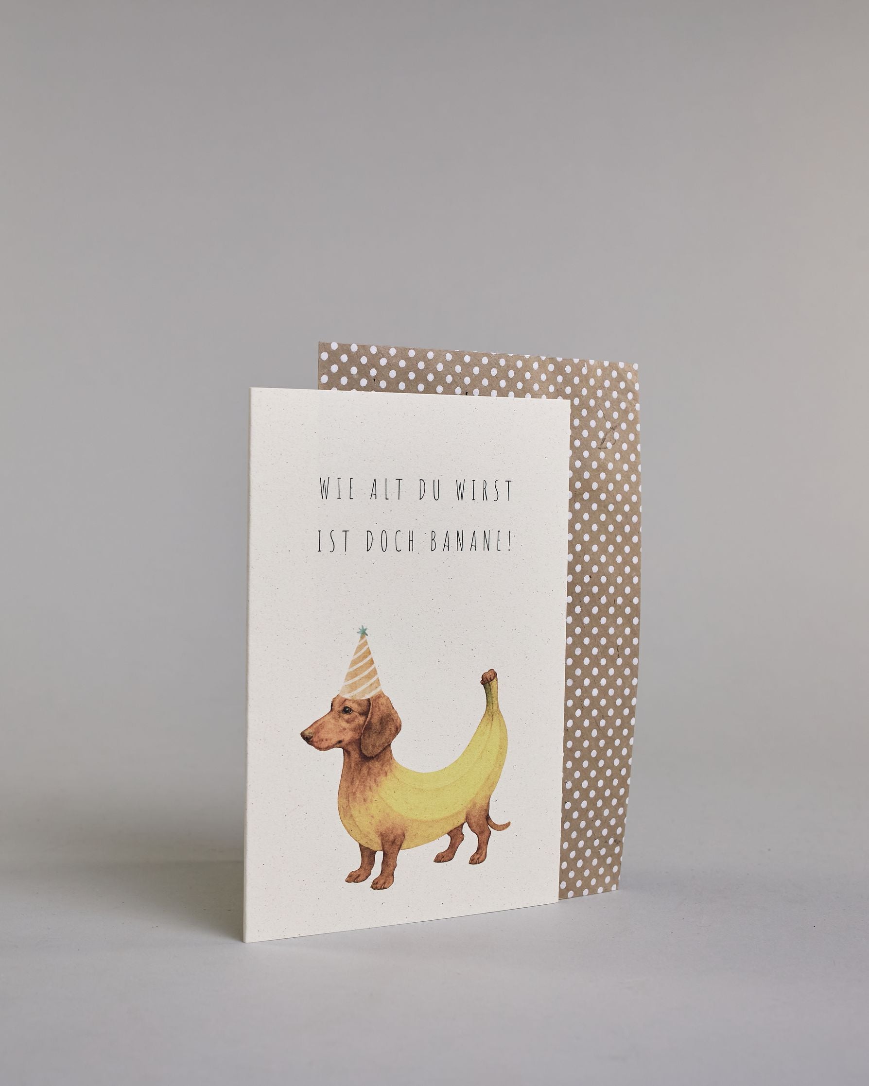 Glückwunschkarte zum Geburtstag mit schwarzer Kalligrafieaufschrift "Wie alt du wirst ist doch Banane!", mit einem genzeichnetem brauner Dackel dessen Körper aussieht wie eine gelbe Banane und einem gelb-orangen Partyhut auf dem Kopf auf hellem Hintergund. Oben steht in zwei Zeilen "WIE ALT DU WIRST", darunter "IST DOCH BANANE!" beides in dünner gerader Kalligrafieschrift und darunter mittig der Bananenhund. Dazu ein beige-weißes handgeschöpftes Kuvert. #3