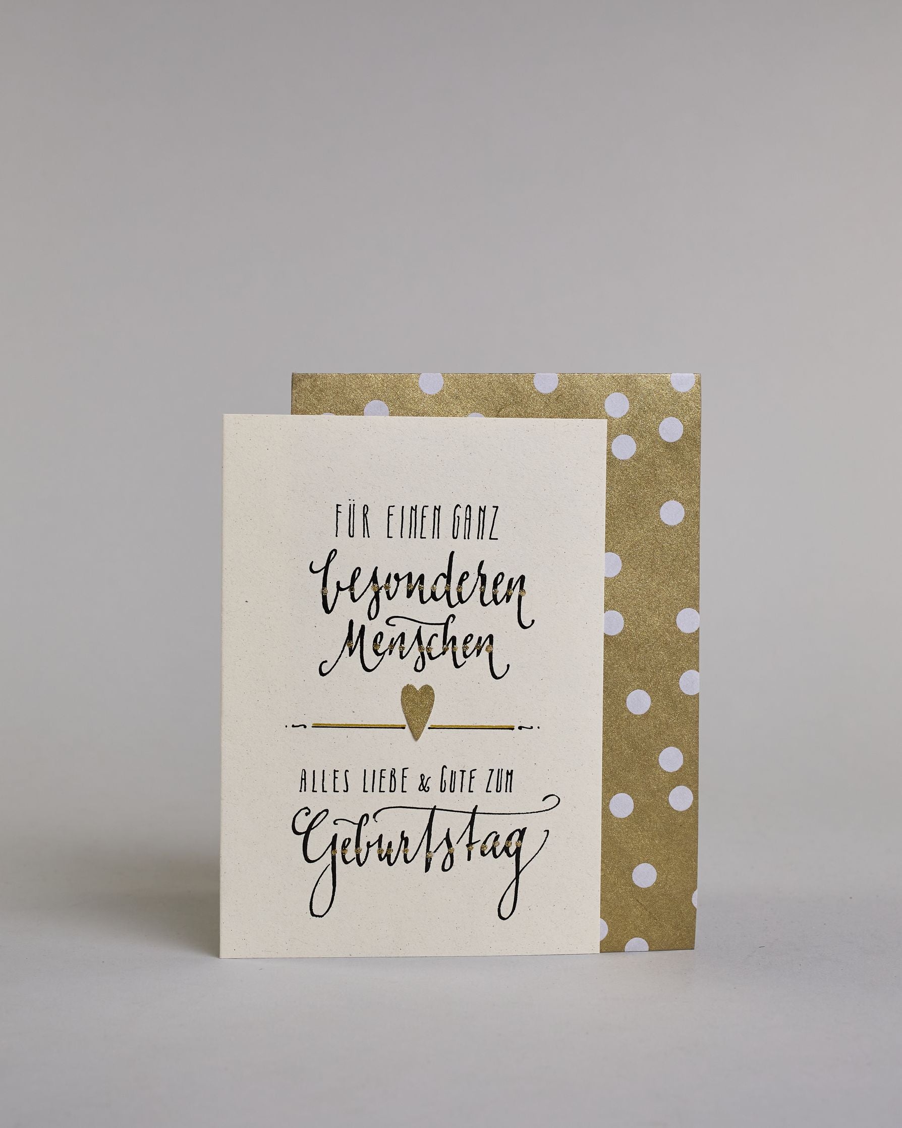Glückwunschkarte zum Geburtstag mit Schwarzer Kalligrafieaufschrift "Für einen ganz besonderen Menschen Alles Liebe & Gute zum Geburtstag" auf hellem Hintergund. Aufgeteilt ist die Schrift wiefolgt: "FÜR EINEN GANZ" in dünner gerader Kalligrafieschrift, darunter "besonderen", darunter "Menschen beides in einer geschwungenen Schreibschrift, darunter mittig ein goldenen aufgeklebtes kleine Herz das links und rechts von horizonatlen Linien umgeben ist, darunter "ALLES LIEBE & GUTE ZUM" in dünner gerade Kallig…