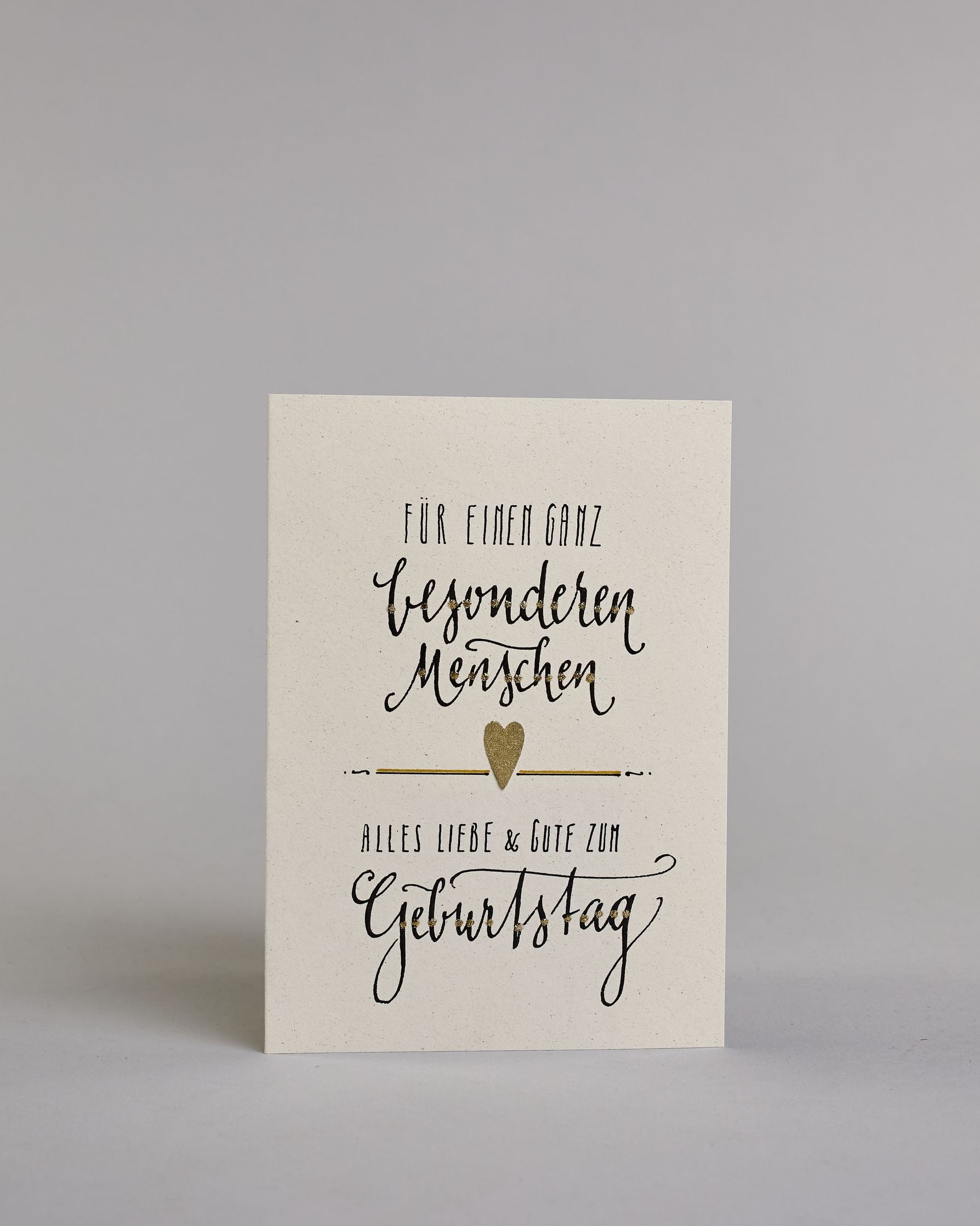 Glückwunschkarte zum Geburtstag mit Schwarzer Kalligrafieaufschrift "Für einen ganz besonderen Menschen Alles Liebe & Gute zum Geburtstag" auf hellem Hintergund. Aufgeteilt ist die Schrift wiefolgt: "FÜR EINEN GANZ" in dünner gerader Kalligrafieschrift, darunter "besonderen", darunter "Menschen beides in einer geschwungenen Schreibschrift, darunter mittig ein goldenen aufgeklebtes kleine Herz das links und rechts von horizonatlen Linien umgeben ist, darunter "ALLES LIEBE & GUTE ZUM" in dünner gerade Kallig…