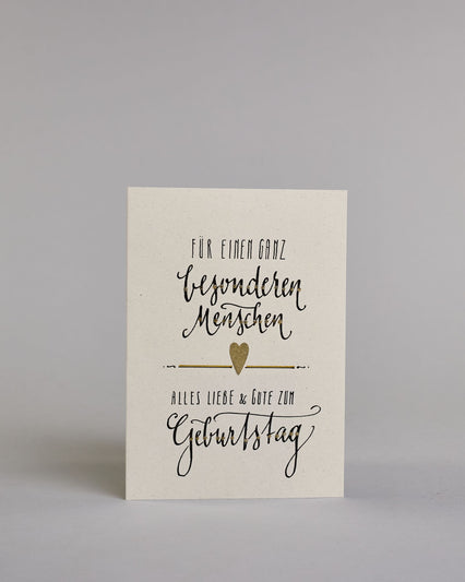 Glückwunschkarte zum Geburtstag mit Schwarzer Kalligrafieaufschrift "Für einen ganz besonderen Menschen Alles Liebe & Gute zum Geburtstag" auf hellem Hintergund. Aufgeteilt ist die Schrift wiefolgt: "FÜR EINEN GANZ" in dünner gerader Kalligrafieschrift, darunter "besonderen", darunter "Menschen beides in einer geschwungenen Schreibschrift, darunter mittig ein goldenen aufgeklebtes kleine Herz das links und rechts von horizonatlen Linien umgeben ist, darunter "ALLES LIEBE & GUTE ZUM" in dünner gerade Kallig…