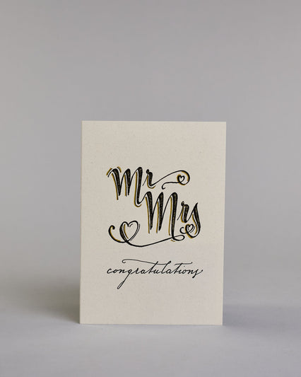 Glückwunschkarte zur Hochzeit in Enlisch mit der Kalligrafieaufschrift "MrMrs congratulations" In großer, geschwungener Schrift mit schwarzen Buchstaben und goldfarbenen Datails steht „Mr Mrs“. Um die Wörter herum sind drei kleine Herzen gezeichnet – eines links unten, eines rechts oben am „r“ und eines rechts unten beim „s“.
Darunter, in kleinerer, feiner Schreibschrift, steht das Wort „congratulations“ in Schwarz.  Dazu ein gold-weißes handgeschöpftes Kuvert. #1