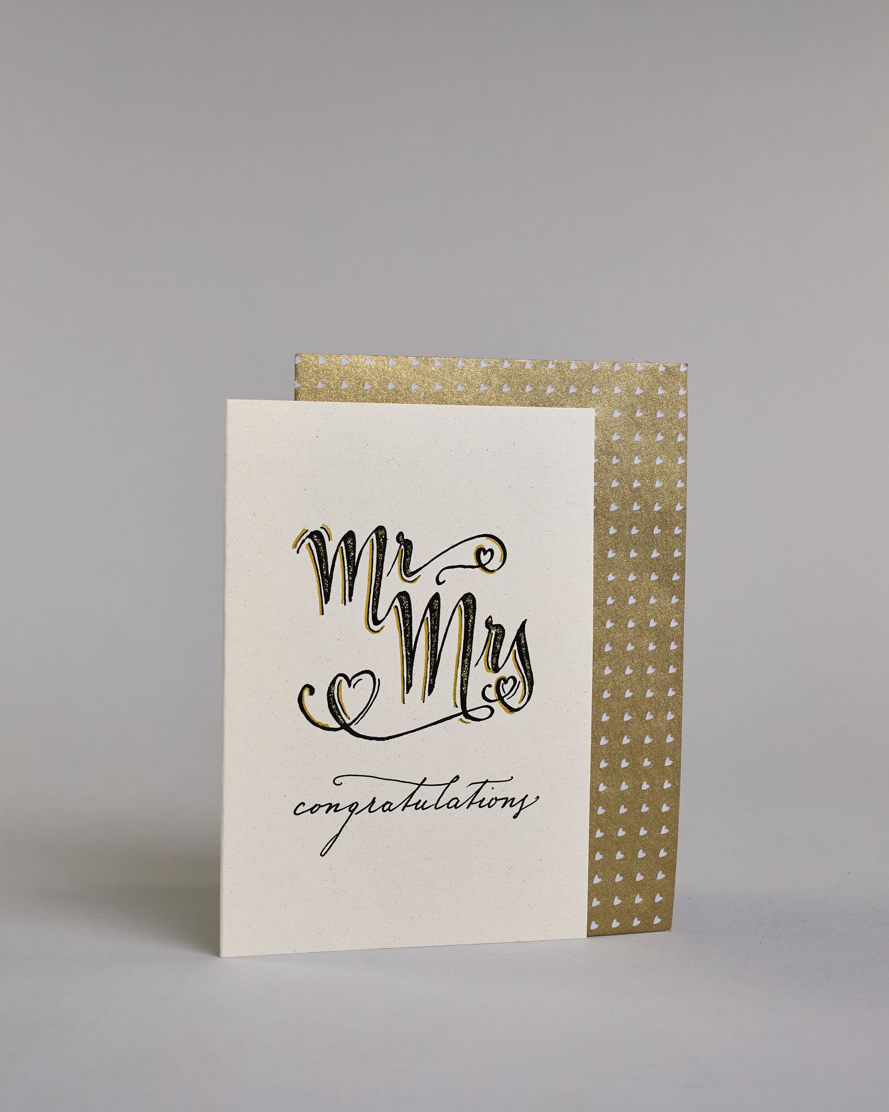 Glückwunschkarte zur Hochzeit in Enlisch mit der Kalligrafieaufschrift "MrMrs congratulations" In großer, geschwungener Schrift mit schwarzen Buchstaben und goldfarbenen Datails steht „Mr Mrs“. Um die Wörter herum sind drei kleine Herzen gezeichnet – eines links unten, eines rechts oben am „r“ und eines rechts unten beim „s“.
Darunter, in kleinerer, feiner Schreibschrift, steht das Wort „congratulations“ in Schwarz.  Dazu ein gold-weißes handgeschöpftes Kuvert. #3