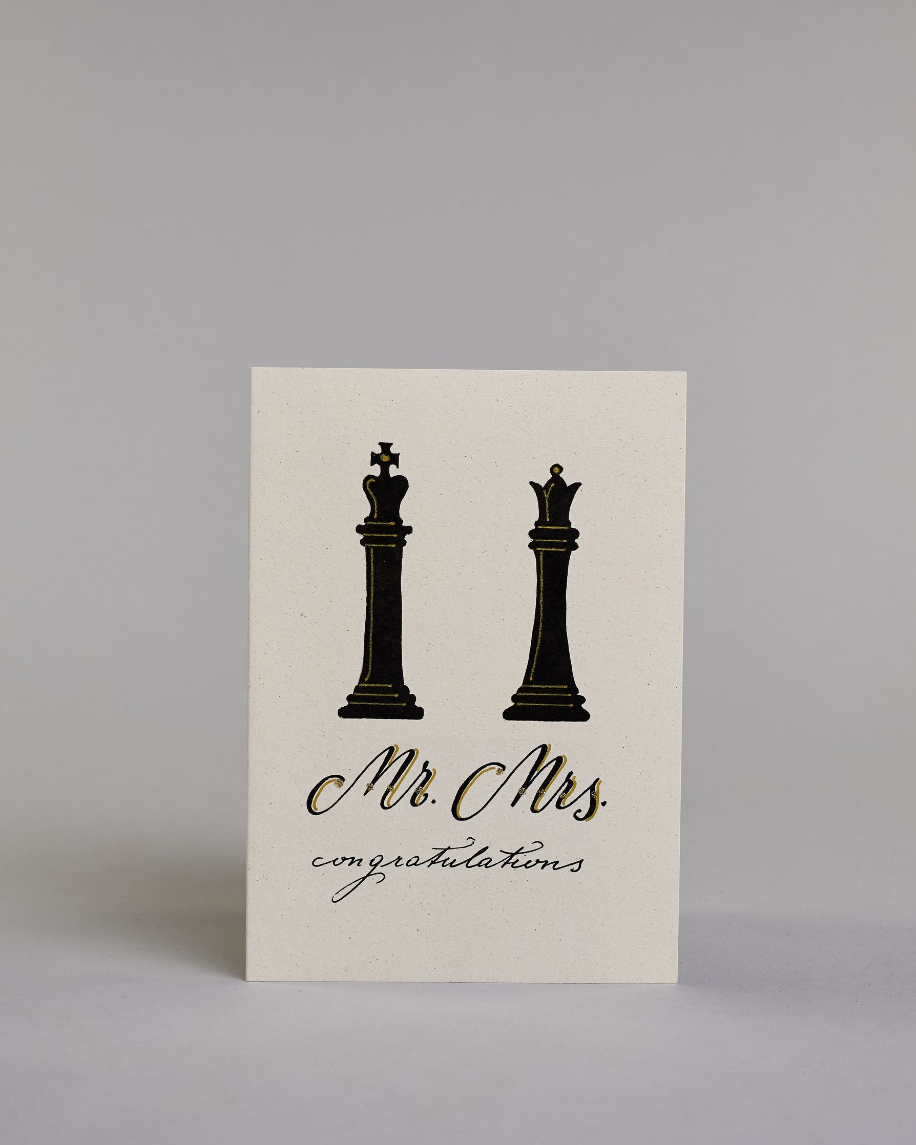 Glückwunschkarte zur Hochzeit in Enlisch mit der Kalligrafieaufschrift "Mr. Mrs. congratulations" auf hellem Hintergrund. Oben sind zwei große Schachfiguren abgebildet: links ein schwarzer König, rechts eine schwarze Dame. Beide Figuren sind mit feinen goldfarbenen Linien akzentuiert.
Darunter steht in geschwungener Schrift mit schwarzen Buchstaben und goldfarbenen Schattierungen „Mr. Mrs.“.
Unterhalb davon folgt in kleiner, feiner schwarzer Schreibschrift das Wort „congratulations“.  Dazu ein gold-weißes…