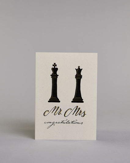 Glückwunschkarte zur Hochzeit in Enlisch mit der Kalligrafieaufschrift "Mr. Mrs. congratulations" auf hellem Hintergrund. Oben sind zwei große Schachfiguren abgebildet: links ein schwarzer König, rechts eine schwarze Dame. Beide Figuren sind mit feinen goldfarbenen Linien akzentuiert.
Darunter steht in geschwungener Schrift mit schwarzen Buchstaben und goldfarbenen Schattierungen „Mr. Mrs.“.
Unterhalb davon folgt in kleiner, feiner schwarzer Schreibschrift das Wort „congratulations“.  Dazu ein gold-weißes…
