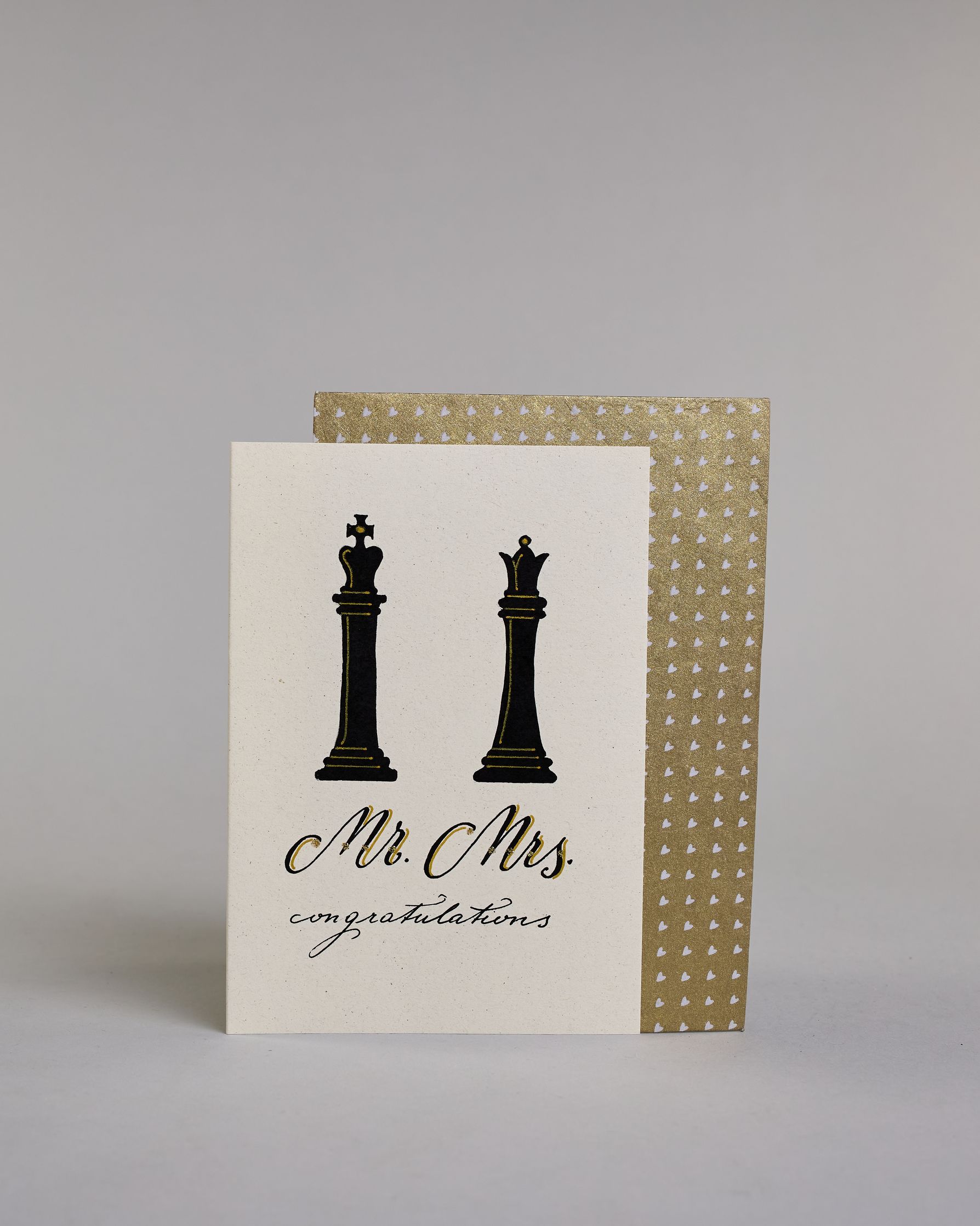 Glückwunschkarte zur Hochzeit in Enlisch mit der Kalligrafieaufschrift "Mr. Mrs. congratulations" auf hellem Hintergrund. Oben sind zwei große Schachfiguren abgebildet: links ein schwarzer König, rechts eine schwarze Dame. Beide Figuren sind mit feinen goldfarbenen Linien akzentuiert.
Darunter steht in geschwungener Schrift mit schwarzen Buchstaben und goldfarbenen Schattierungen „Mr. Mrs.“.
Unterhalb davon folgt in kleiner, feiner schwarzer Schreibschrift das Wort „congratulations“.  Dazu ein gold-weißes…