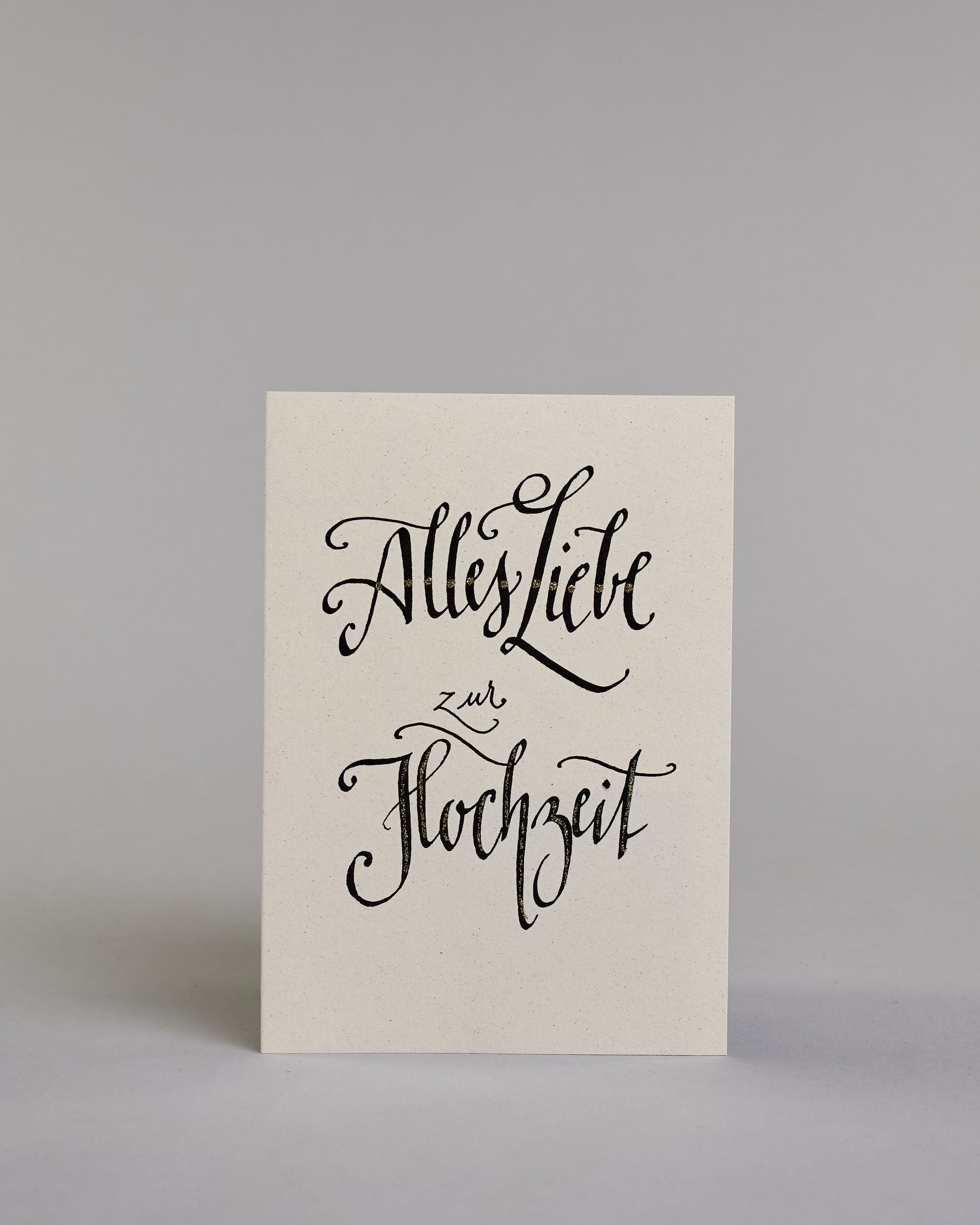 Glückwunschkarte zur Hochzeit mit der Kalligrafieaufschrift "Alles Liebe zur Hochzeit" auf hellem Hintergrund. Die Schrift ist mit feinen goldfarbenen Akzenten versehen. Das Wort „zur“ ist kleiner gesetzt und steht zwischen den beiden großen Wörtern „Liebe“ und „Hochzeit“. Dazu ein gold-weißes handgeschöpftes Kuvert. #1