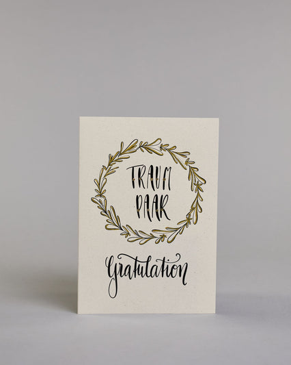 Glückwunschkarte zur Hochzeit mit der Kalligrafieaufschrift "Traumpaar Gratulation" auf hellem Hintergrund. In der Mitte eines runden, handgezeichneten Blätterkranzes, der mit goldfarbenen Akzenten versehen ist, steht in schwarzer Schrift das Wort „TRAUM PAAR“ auf zwei Zeilen. Darunter steht in größerer, geschwungener schwarzer Schrift das Wort „Gratulation“. Dazu ein gold-weißes handgeschöpftes Kuvert. #1