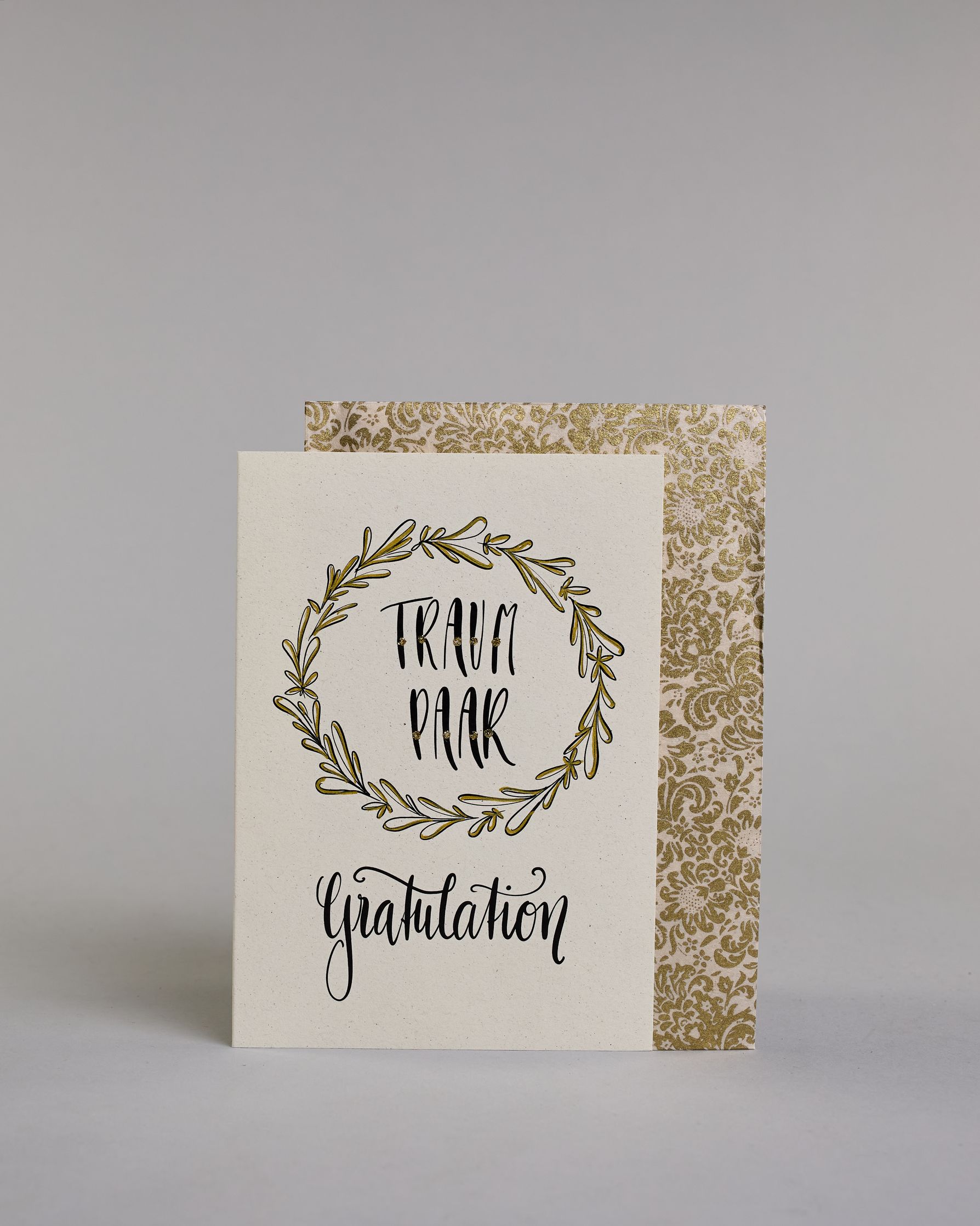Glückwunschkarte zur Hochzeit mit der Kalligrafieaufschrift "Traumpaar Gratulation" auf hellem Hintergrund. In der Mitte eines runden, handgezeichneten Blätterkranzes, der mit goldfarbenen Akzenten versehen ist, steht in schwarzer Schrift das Wort „TRAUM PAAR“ auf zwei Zeilen. Darunter steht in größerer, geschwungener schwarzer Schrift das Wort „Gratulation“. Dazu ein gold-weißes handgeschöpftes Kuvert. #2