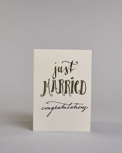 Glückwunschkarte zur Hochzeit in Englisch mit der Kalligrafieaufschrift "just MARRIED congratulations" auf hellem Hintergrund. Oben in schwungvoller schwarzer Schrift steht das Wort „just“. Darunter in größerer, dekorativer Schrift mit goldfarbenen Akzenten das Wort „MARRIED“. Der Punkt vom „I“ ist ein kleines Herz. Am unteren Rand der Karte steht in eleganter, geschwungener schwarzer Schrift „congratulations“. Dazu ein gold-weißes handgeschöpftes Kuvert. #1