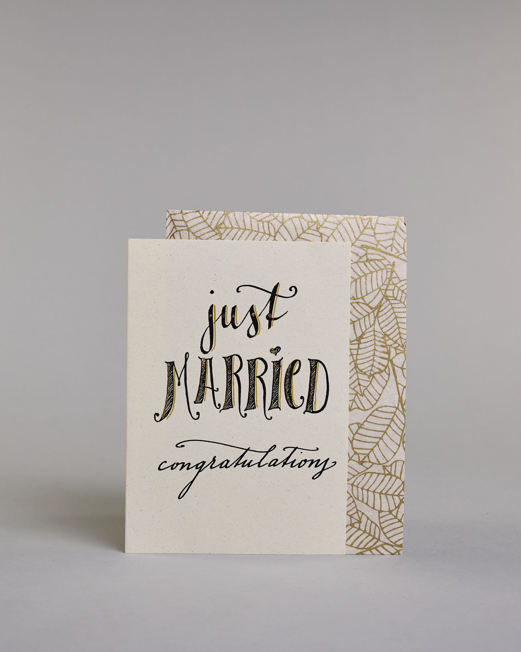 Glückwunschkarte zur Hochzeit in Englisch mit der Kalligrafieaufschrift "just MARRIED congratulations" auf hellem Hintergrund. Oben in schwungvoller schwarzer Schrift steht das Wort „just“. Darunter in größerer, dekorativer Schrift mit goldfarbenen Akzenten das Wort „MARRIED“. Der Punkt vom „I“ ist ein kleines Herz. Am unteren Rand der Karte steht in eleganter, geschwungener schwarzer Schrift „congratulations“. Dazu ein gold-weißes handgeschöpftes Kuvert. #2