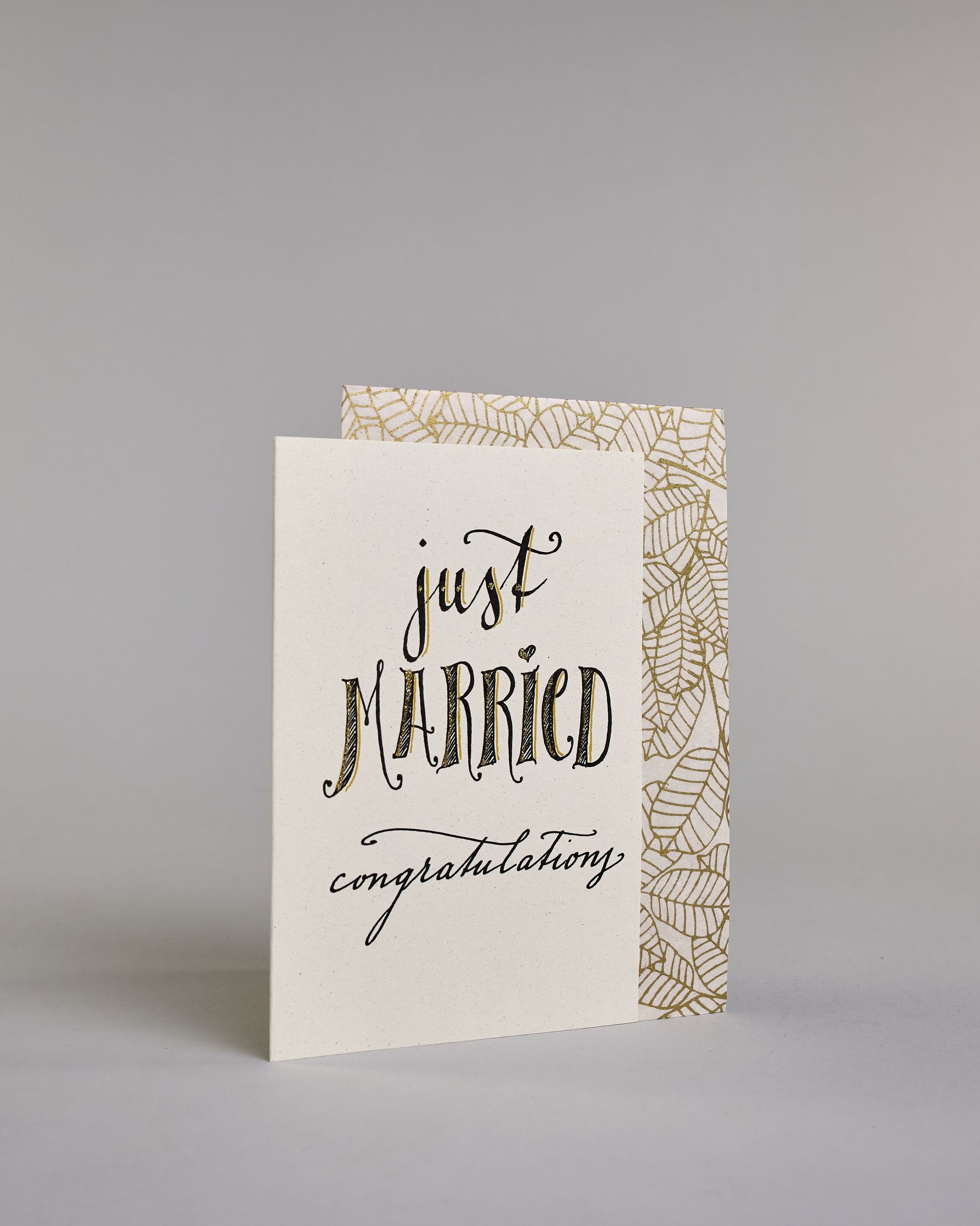 Glückwunschkarte zur Hochzeit in Englisch mit der Kalligrafieaufschrift "just MARRIED congratulations" auf hellem Hintergrund. Oben in schwungvoller schwarzer Schrift steht das Wort „just“. Darunter in größerer, dekorativer Schrift mit goldfarbenen Akzenten das Wort „MARRIED“. Der Punkt vom „I“ ist ein kleines Herz. Am unteren Rand der Karte steht in eleganter, geschwungener schwarzer Schrift „congratulations“. Dazu ein gold-weißes handgeschöpftes Kuvert. #3