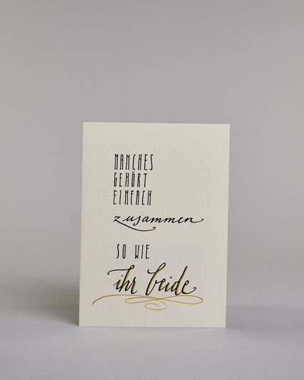 Glückwunschkarte zur Hochzeit mit der Kalligrafieaufschrift "Manchses gehört einfach zusammen so wie ihr beide" auf einem hellen Hintergrund. Sie zeigt den Text in verschiedenen Schriften und Größen: Oben steht in schmaler, schwarzer Druckschrift „MANCHES GEHÖRT EINFACH“, darunter in schwungvoller, kleinerer Schreibschrift „zusammen“, darunter wieder in schmaler Druckschrift „SO WIE“ und am unteren Rand in großer, geschwungener Schrift mit goldfarbenen Akzenten „ihr beide“, unterstrichen von einem dekorati…