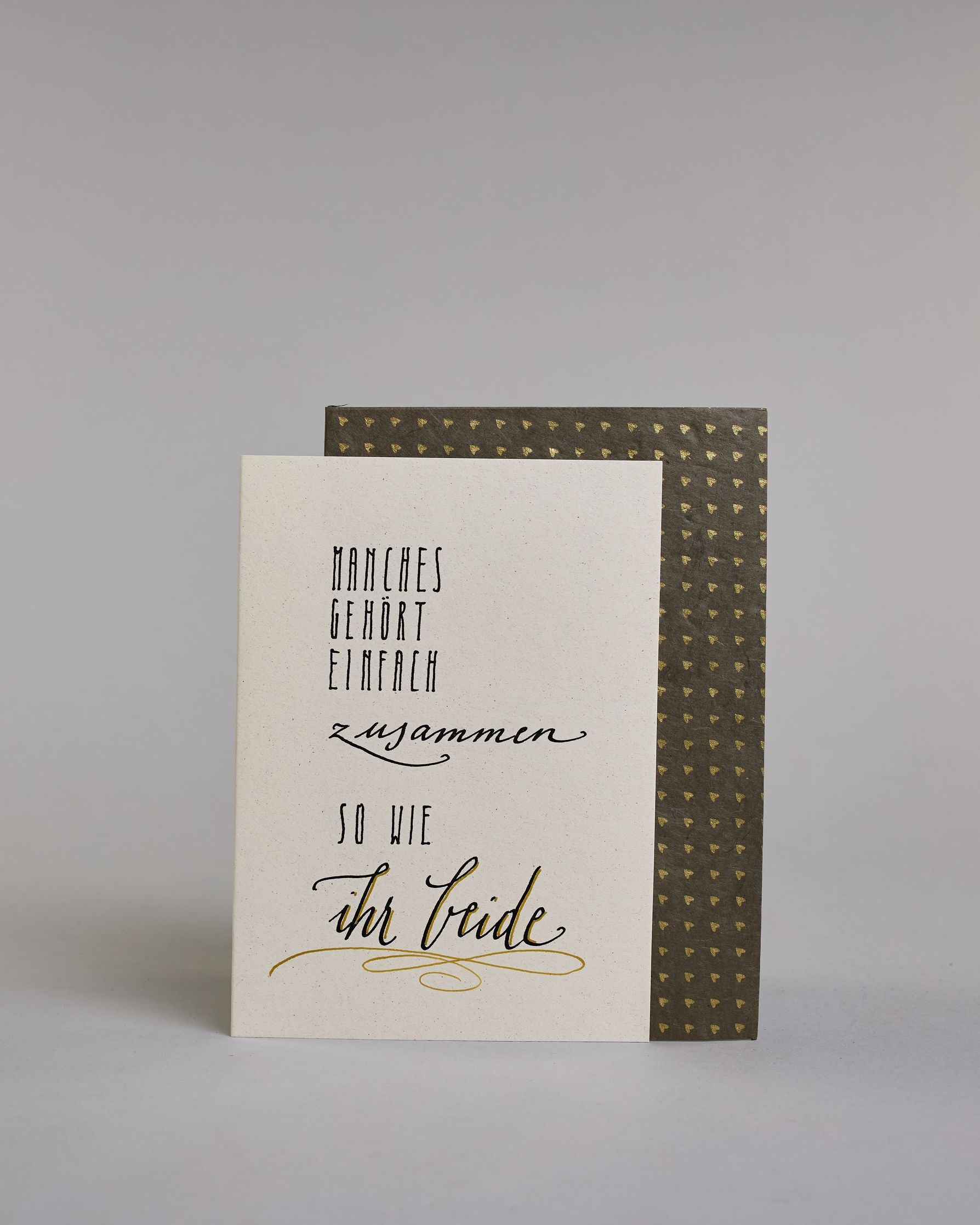 Glückwunschkarte zur Hochzeit mit der Kalligrafieaufschrift "Manchses gehört einfach zusammen so wie ihr beide" auf einem hellen Hintergrund. Sie zeigt den Text in verschiedenen Schriften und Größen: Oben steht in schmaler, schwarzer Druckschrift „MANCHES GEHÖRT EINFACH“, darunter in schwungvoller, kleinerer Schreibschrift „zusammen“, darunter wieder in schmaler Druckschrift „SO WIE“ und am unteren Rand in großer, geschwungener Schrift mit goldfarbenen Akzenten „ihr beide“, unterstrichen von einem dekorati…