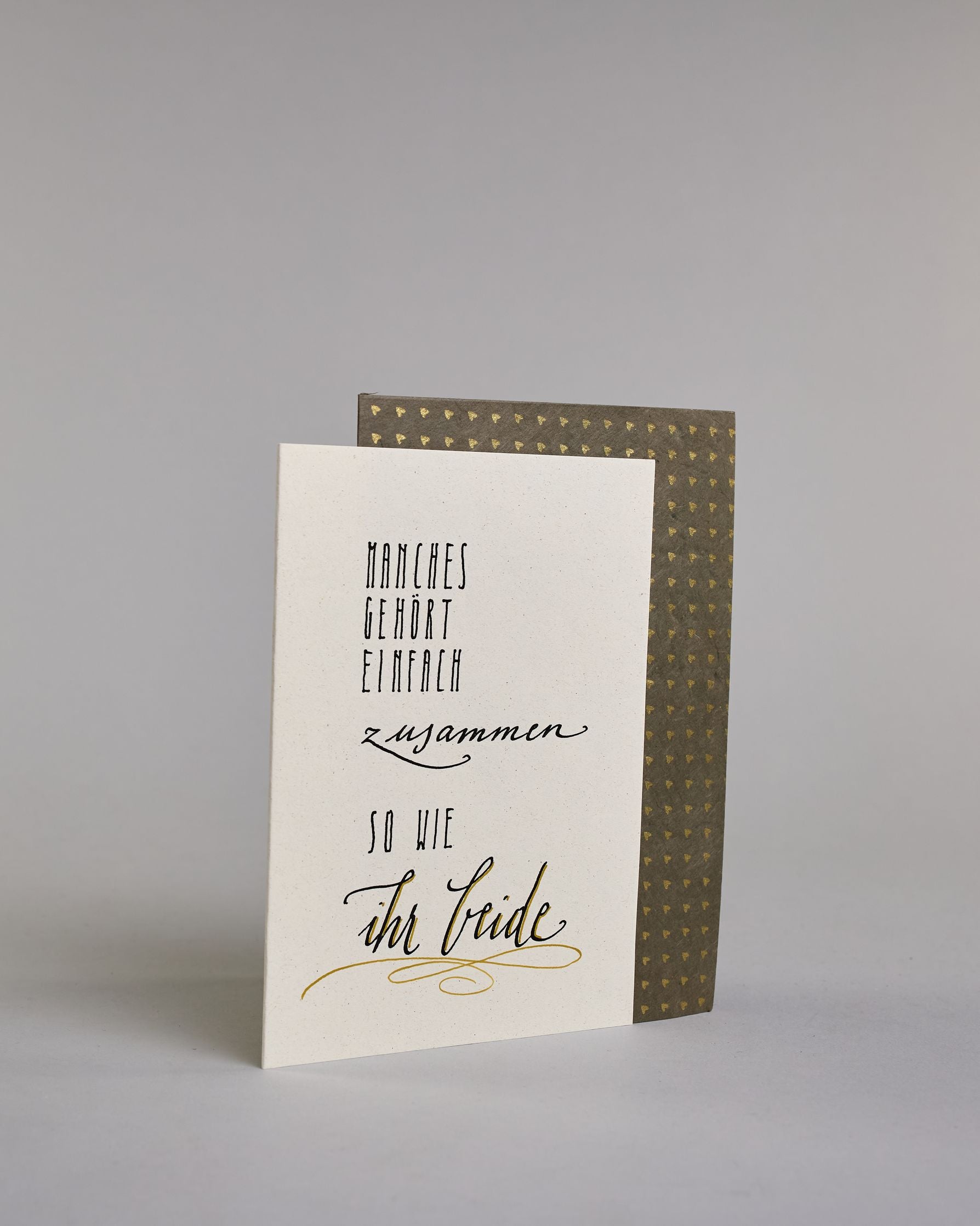 Glückwunschkarte zur Hochzeit mit der Kalligrafieaufschrift "Manchses gehört einfach zusammen so wie ihr beide" auf einem hellen Hintergrund. Sie zeigt den Text in verschiedenen Schriften und Größen: Oben steht in schmaler, schwarzer Druckschrift „MANCHES GEHÖRT EINFACH“, darunter in schwungvoller, kleinerer Schreibschrift „zusammen“, darunter wieder in schmaler Druckschrift „SO WIE“ und am unteren Rand in großer, geschwungener Schrift mit goldfarbenen Akzenten „ihr beide“, unterstrichen von einem dekorati…