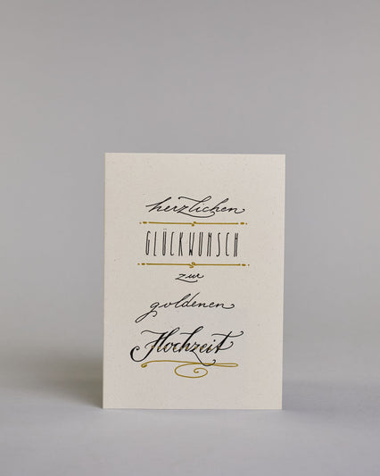 Glückwunschkarte zur Hochzeit mit der Kalligrafieaufschrift "herzlichen Glückwunsch zur goldenen Hochzeit" auf hellem Hintergrund. Sie zeigt den Text in unterschiedlichen Schriften: Oben steht in kleiner schwarzer Schreibschrift „herzlichen“, darunter in schmaler schwarzer Druckschrift das Wort „GLÜCKWUNSCH“, eingerahmt von goldenen Punkten und Linien, darunter in kleiner schwarzer Schreibschrift das Wort „zur“, darunter in schwarzer Schreibschrift mit schwungvollen Buchstaben „goldenen“, und am unteren Ra…