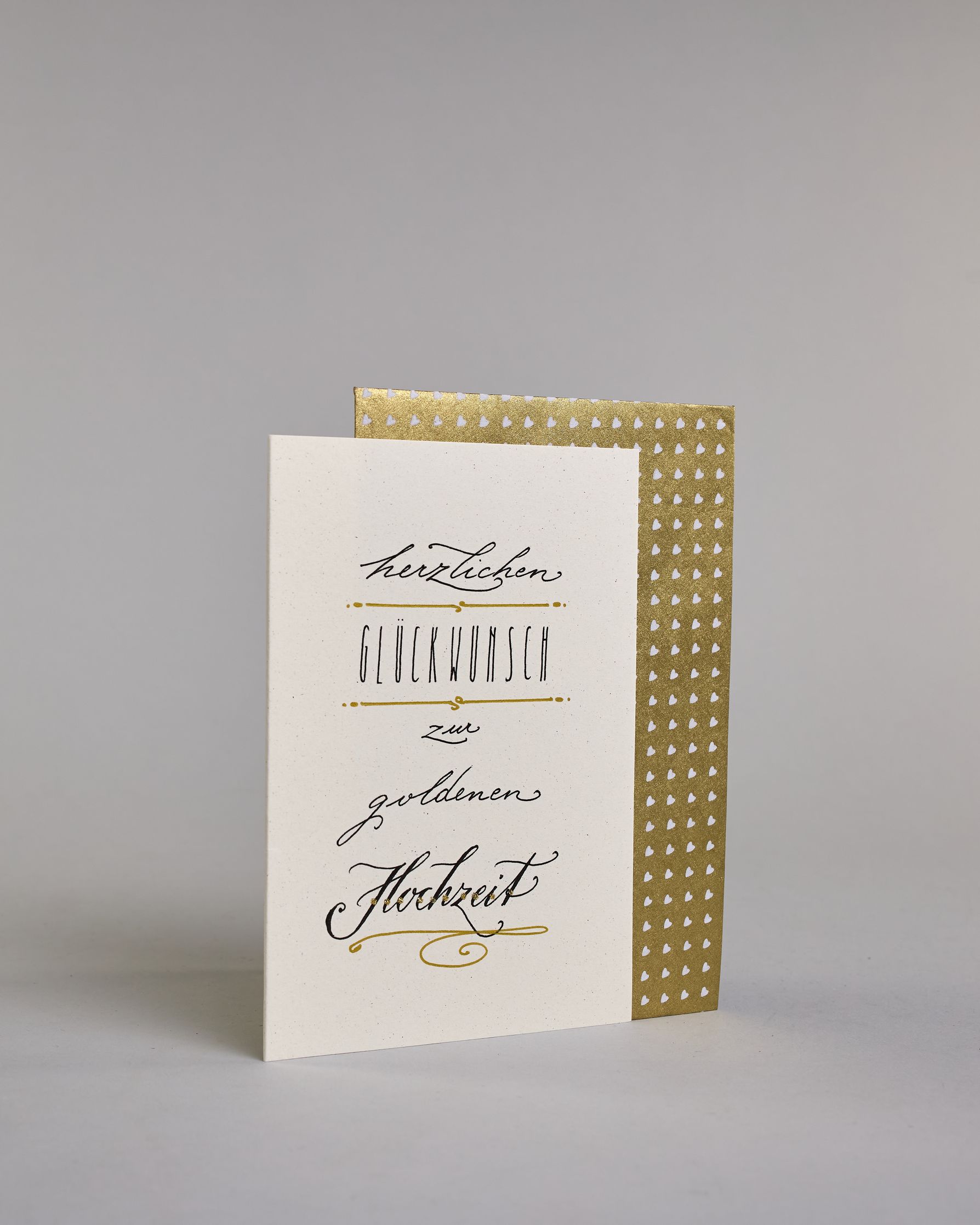 Glückwunschkarte zur Hochzeit mit der Kalligrafieaufschrift "herzlichen Glückwunsch zur goldenen Hochzeit" auf hellem Hintergrund. Sie zeigt den Text in unterschiedlichen Schriften: Oben steht in kleiner schwarzer Schreibschrift „herzlichen“, darunter in schmaler schwarzer Druckschrift das Wort „GLÜCKWUNSCH“, eingerahmt von goldenen Punkten und Linien, darunter in kleiner schwarzer Schreibschrift das Wort „zur“, darunter in schwarzer Schreibschrift mit schwungvollen Buchstaben „goldenen“, und am unteren Ra…