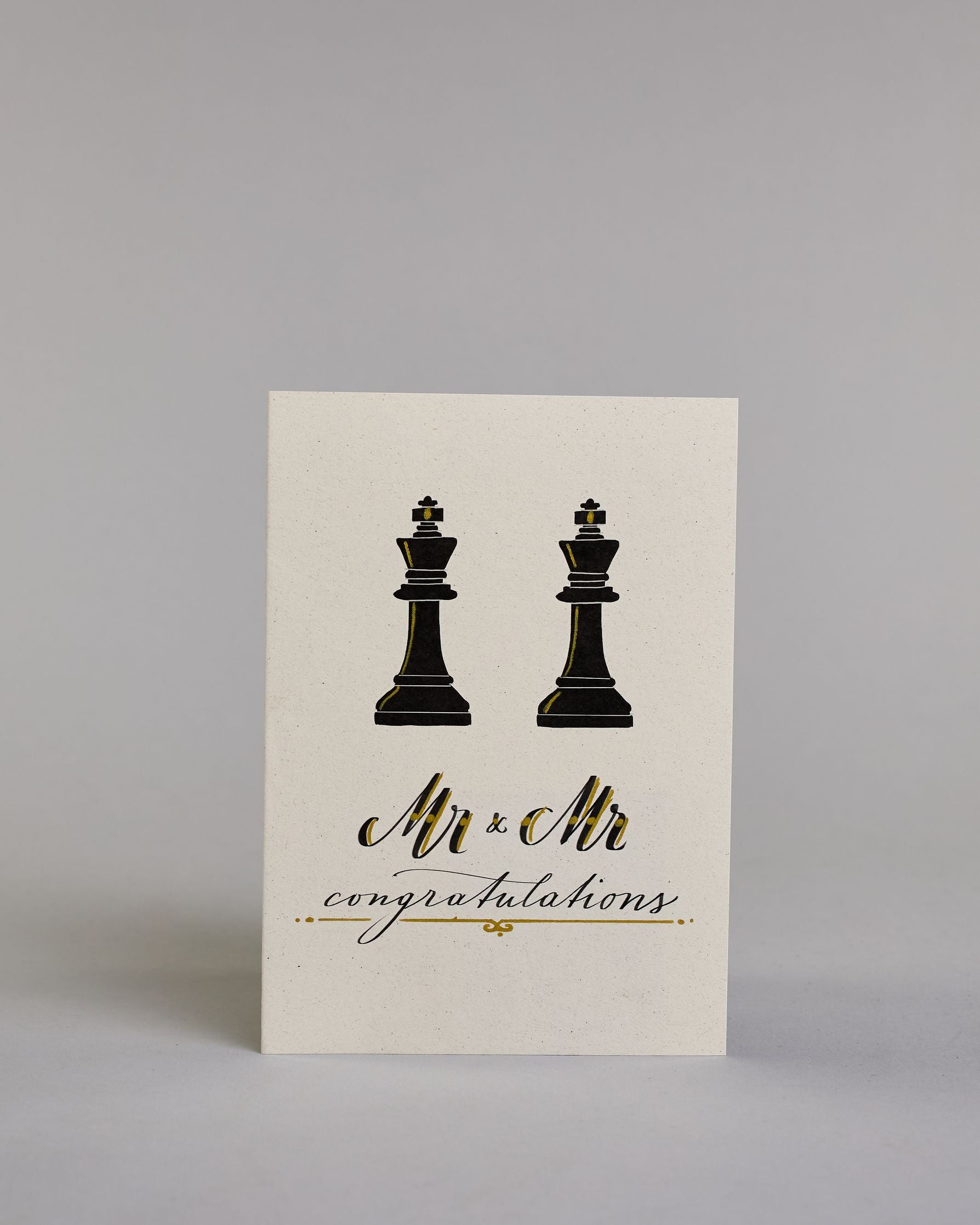 Glückwunschkarte zur Hochzeit in Englisch für zwei Männer mit der Kalligrafieaufschrift "Mr & Mr congratulations" auf hellem Hintergrund. Sie zeigt oben zwei schwarze Schachfiguren in Form von Königen, die mit feinen goldenen Linien akzentuiert sind. Darunter steht in großer, geschwungener schwarzer Schrift mit goldenen Details „Mr & Mr“. Unterhalb davon befindet sich in kleinerer schwarzer Schreibschrift das Wort „congratulations“, eingerahmt von zwei kleinen goldenen Punkten und unterstrichen mit einer g…