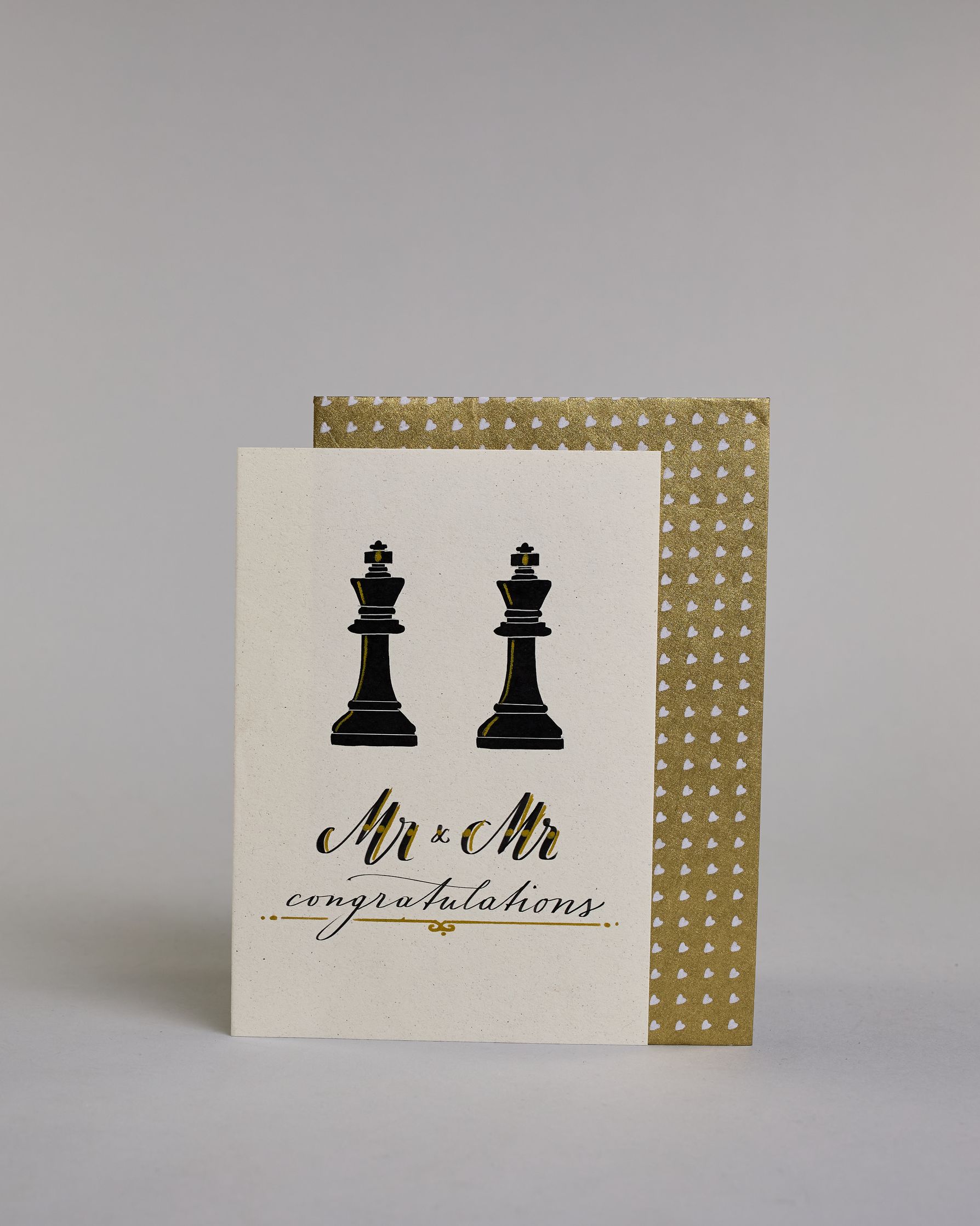 Glückwunschkarte zur Hochzeit in Englisch für zwei Männer mit der Kalligrafieaufschrift "Mr & Mr congratulations" auf hellem Hintergrund. Sie zeigt oben zwei schwarze Schachfiguren in Form von Königen, die mit feinen goldenen Linien akzentuiert sind. Darunter steht in großer, geschwungener schwarzer Schrift mit goldenen Details „Mr & Mr“. Unterhalb davon befindet sich in kleinerer schwarzer Schreibschrift das Wort „congratulations“, eingerahmt von zwei kleinen goldenen Punkten und unterstrichen mit einer g…