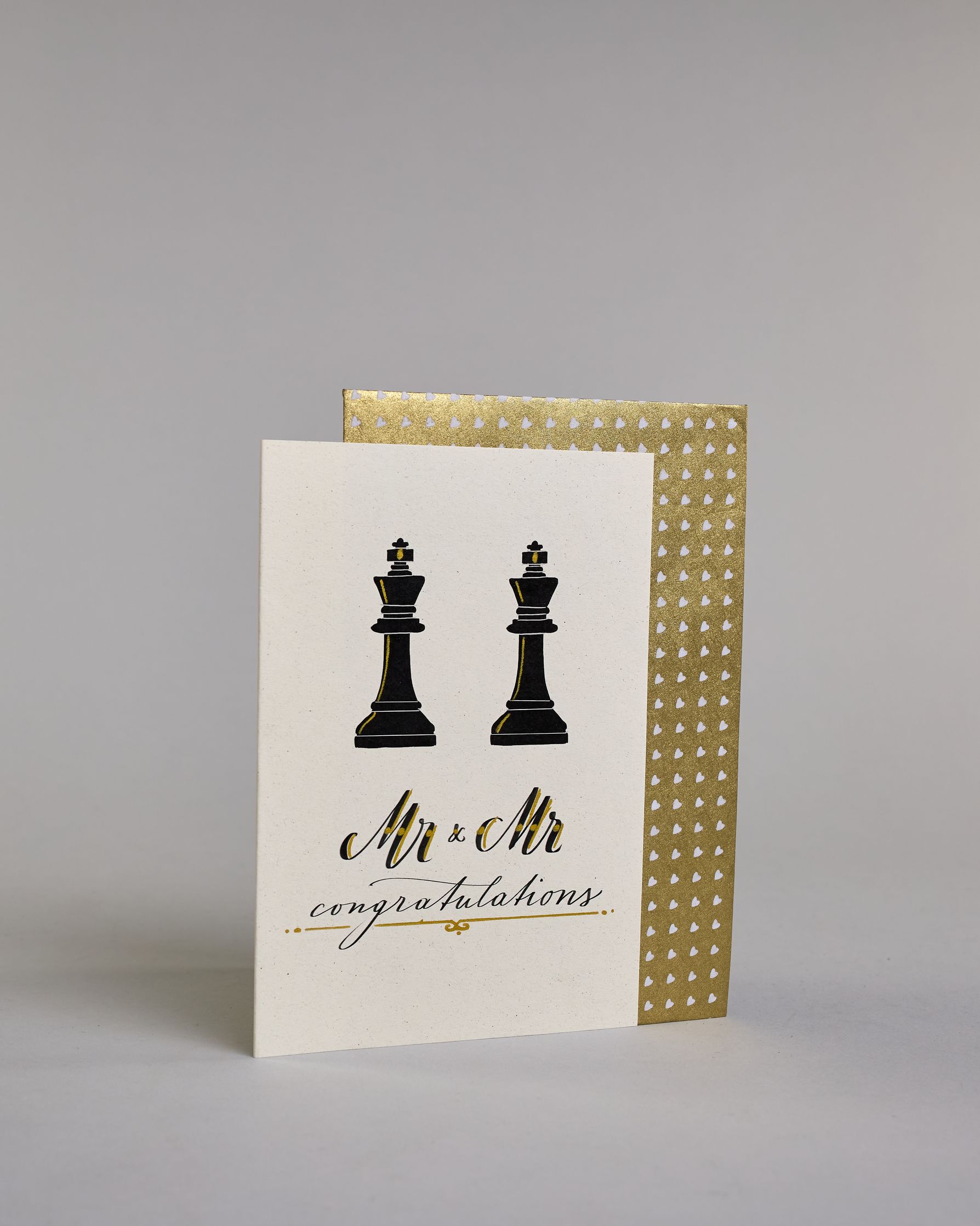 Glückwunschkarte zur Hochzeit in Englisch für zwei Männer mit der Kalligrafieaufschrift "Mr & Mr congratulations" auf hellem Hintergrund. Sie zeigt oben zwei schwarze Schachfiguren in Form von Königen, die mit feinen goldenen Linien akzentuiert sind. Darunter steht in großer, geschwungener schwarzer Schrift mit goldenen Details „Mr & Mr“. Unterhalb davon befindet sich in kleinerer schwarzer Schreibschrift das Wort „congratulations“, eingerahmt von zwei kleinen goldenen Punkten und unterstrichen mit einer g…