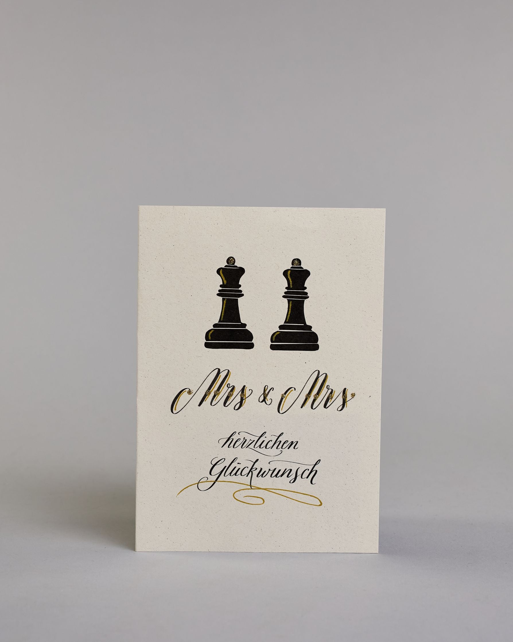Glückwunschkarte zur Hochzeit für zwei Frauen mit der Kalligrafieaufschrift "Mrs & Mrs herzlichen Glückwunsch" auf hellem Hintergrund. Sie zeigt oben zwei schwarze Schachfiguren in Form von Bauern, die mit feinen goldenen Linien akzentuiert sind. Darunter steht in großer, geschwungener schwarzer Schrift mit goldenen Details „Mrs & Mrs“. Unterhalb davon befindet sich in geschwungener schwarzer Schreibschrift auf zwei Zeilen "herzlichen Glückwunsch" darunter ist ein kunstvoller goldener Schwung. Dazu ein gol…