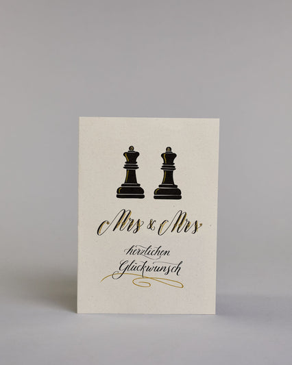 Glückwunschkarte zur Hochzeit für zwei Frauen mit der Kalligrafieaufschrift "Mrs & Mrs herzlichen Glückwunsch" auf hellem Hintergrund. Sie zeigt oben zwei schwarze Schachfiguren in Form von Bauern, die mit feinen goldenen Linien akzentuiert sind. Darunter steht in großer, geschwungener schwarzer Schrift mit goldenen Details „Mrs & Mrs“. Unterhalb davon befindet sich in geschwungener schwarzer Schreibschrift auf zwei Zeilen "herzlichen Glückwunsch" darunter ist ein kunstvoller goldener Schwung. Dazu ein gol…