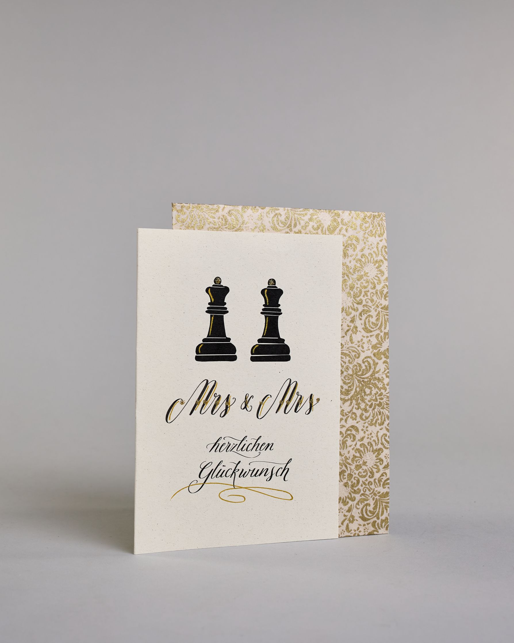Glückwunschkarte zur Hochzeit für zwei Frauen mit der Kalligrafieaufschrift "Mrs & Mrs herzlichen Glückwunsch" auf hellem Hintergrund. Sie zeigt oben zwei schwarze Schachfiguren in Form von Bauern, die mit feinen goldenen Linien akzentuiert sind. Darunter steht in großer, geschwungener schwarzer Schrift mit goldenen Details „Mrs & Mrs“. Unterhalb davon befindet sich in geschwungener schwarzer Schreibschrift auf zwei Zeilen "herzlichen Glückwunsch" darunter ist ein kunstvoller goldener Schwung. Dazu ein gol…