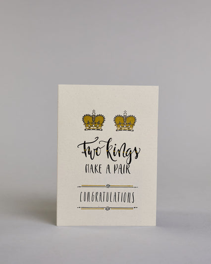 Glückwunschkarte auf Englisch zur Hochzeit für zwei Männer mit der Kalligrafieaufschrift "Two Kings make a pair congratulations" auf hellem Hintergrund. Sie zeigt oben zwei Kronen welche mit Gold verziert sind. Darunter steht in großer, geschwungener schwarzer Schrift mit goldenen Details „Two kings“, darunter mit dünner Kalligraphieschrift "MAKE A PAIR". Unterhalb davon befindet sich in ebenfalls gerader schwarzer Schrift "CONGRATULATIONS" welches sowohl ober- als auch unterhalb prunkvolle horizotale Lini…