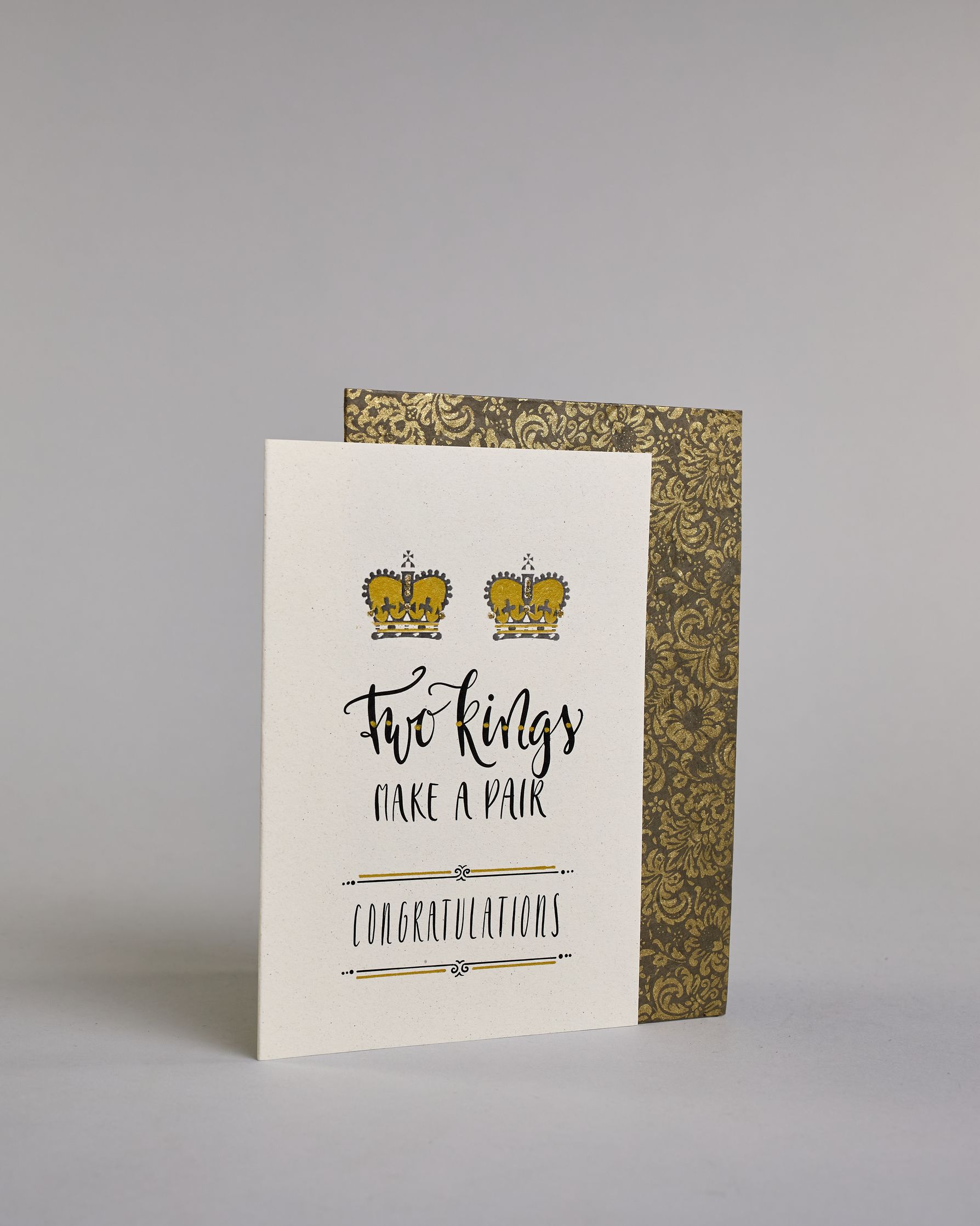 Glückwunschkarte auf Englisch zur Hochzeit für zwei Männer mit der Kalligrafieaufschrift "Two Kings make a pair congratulations" auf hellem Hintergrund. Sie zeigt oben zwei Kronen welche mit Gold verziert sind. Darunter steht in großer, geschwungener schwarzer Schrift mit goldenen Details „Two kings“, darunter mit dünner Kalligraphieschrift "MAKE A PAIR". Unterhalb davon befindet sich in ebenfalls gerader schwarzer Schrift "CONGRATULATIONS" welches sowohl ober- als auch unterhalb prunkvolle horizotale Lini…