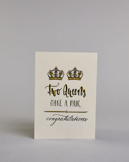 Glückwunschkarte auf Englisch zur Hochzeit für zwei Frauen mit der Kalligrafieaufschrift "Two Queens make a pair congratulations" auf hellem Hintergrund. Sie zeigt oben zwei Kronen welche mit Gold verziert sind. Darunter steht in großer, geschwungener schwarzer Schrift mit goldenen Details „Two Queens“, darunter mit dünner Kalligraphieschrift mit Golddetails "MAKE A PAIR". Unterhalb davon befindet sich ein schwarze prunkvolle horizontale Trennlinie und darunter in geschwungener schwarzer Schreibschrift "co…