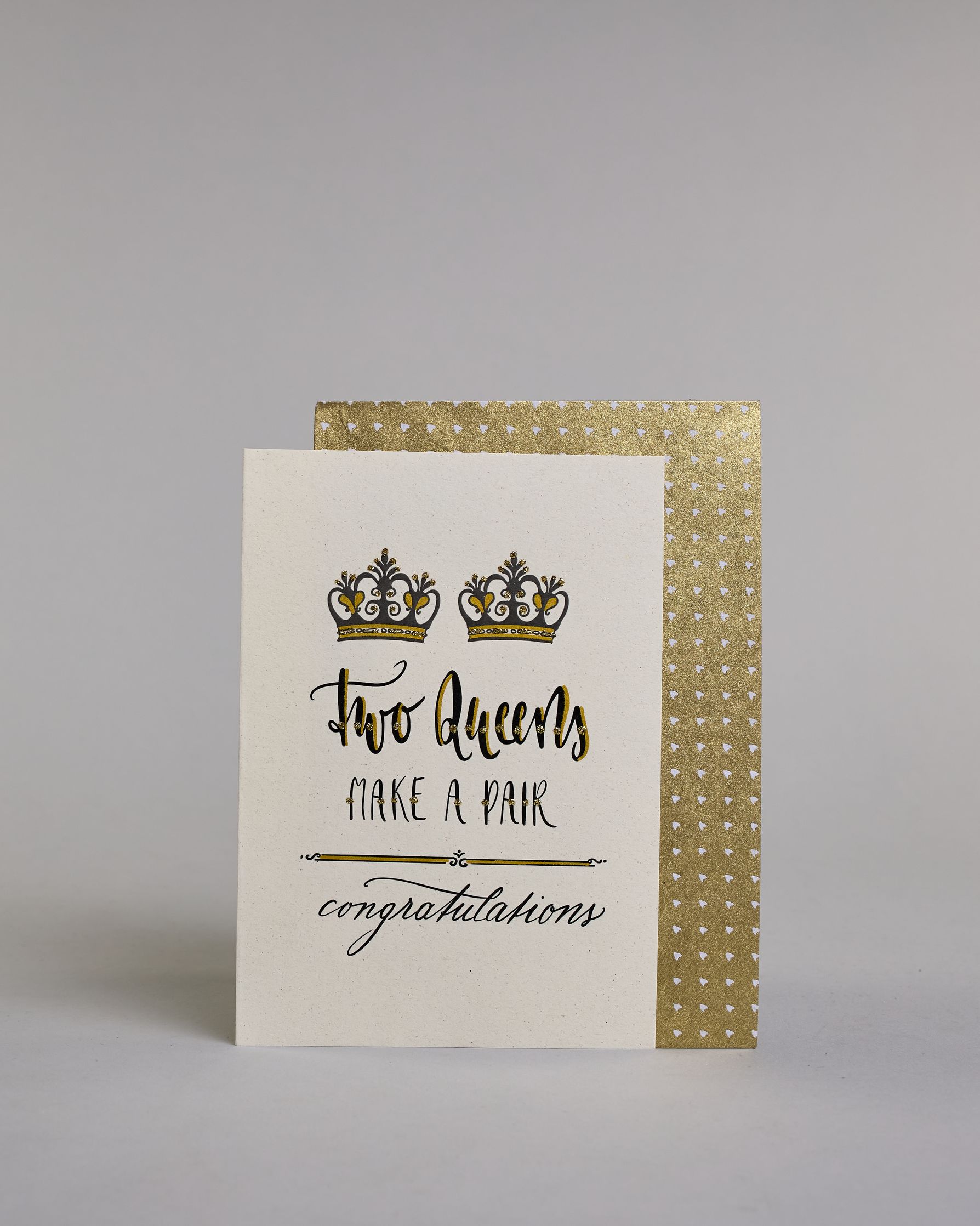 Glückwunschkarte auf Englisch zur Hochzeit für zwei Frauen mit der Kalligrafieaufschrift "Two Queens make a pair congratulations" auf hellem Hintergrund. Sie zeigt oben zwei Kronen welche mit Gold verziert sind. Darunter steht in großer, geschwungener schwarzer Schrift mit goldenen Details „Two Queens“, darunter mit dünner Kalligraphieschrift mit Golddetails "MAKE A PAIR". Unterhalb davon befindet sich ein schwarze prunkvolle horizontale Trennlinie und darunter in geschwungener schwarzer Schreibschrift "co…