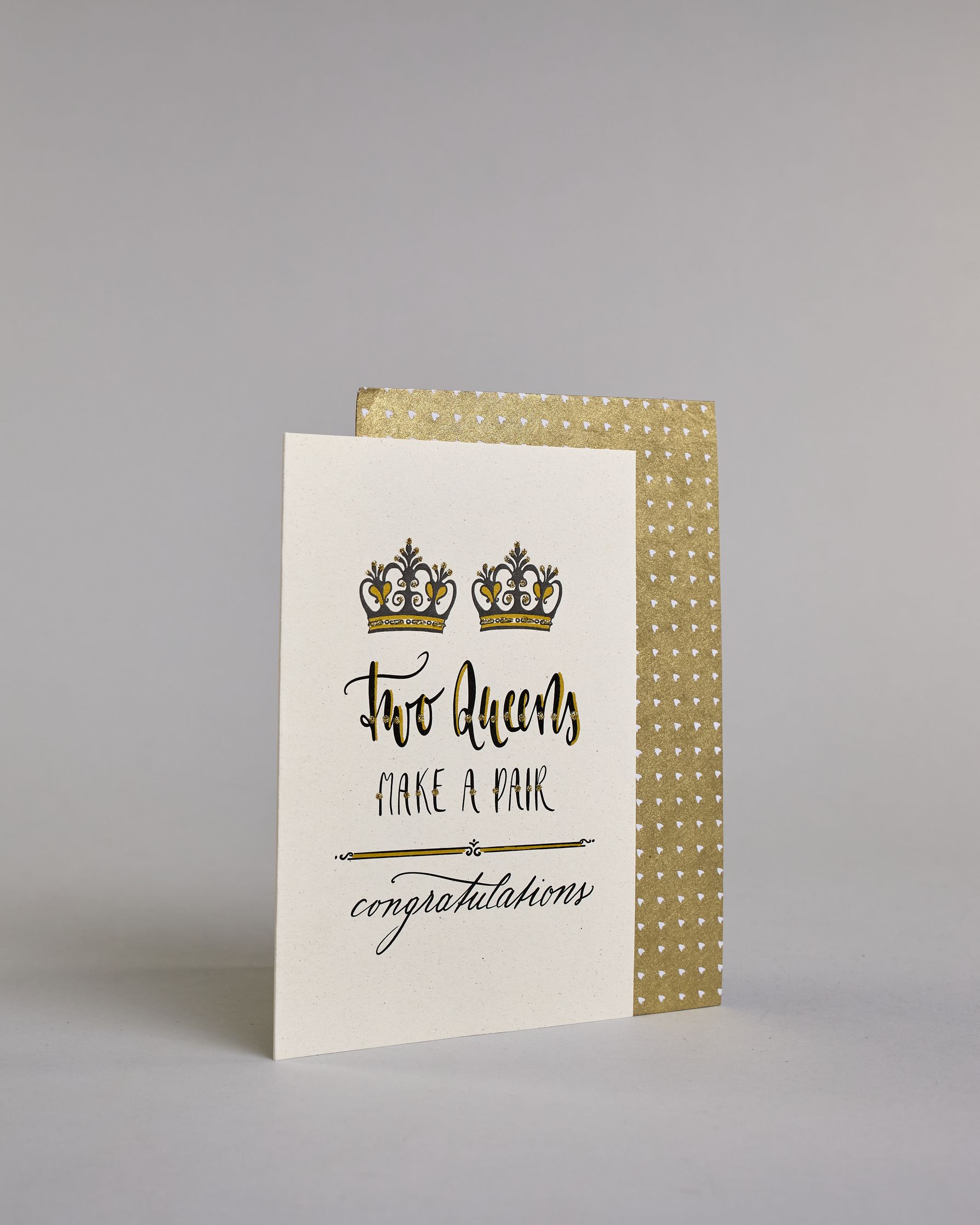 Glückwunschkarte auf Englisch zur Hochzeit für zwei Frauen mit der Kalligrafieaufschrift "Two Queens make a pair congratulations" auf hellem Hintergrund. Sie zeigt oben zwei Kronen welche mit Gold verziert sind. Darunter steht in großer, geschwungener schwarzer Schrift mit goldenen Details „Two Queens“, darunter mit dünner Kalligraphieschrift mit Golddetails "MAKE A PAIR". Unterhalb davon befindet sich ein schwarze prunkvolle horizontale Trennlinie und darunter in geschwungener schwarzer Schreibschrift "co…