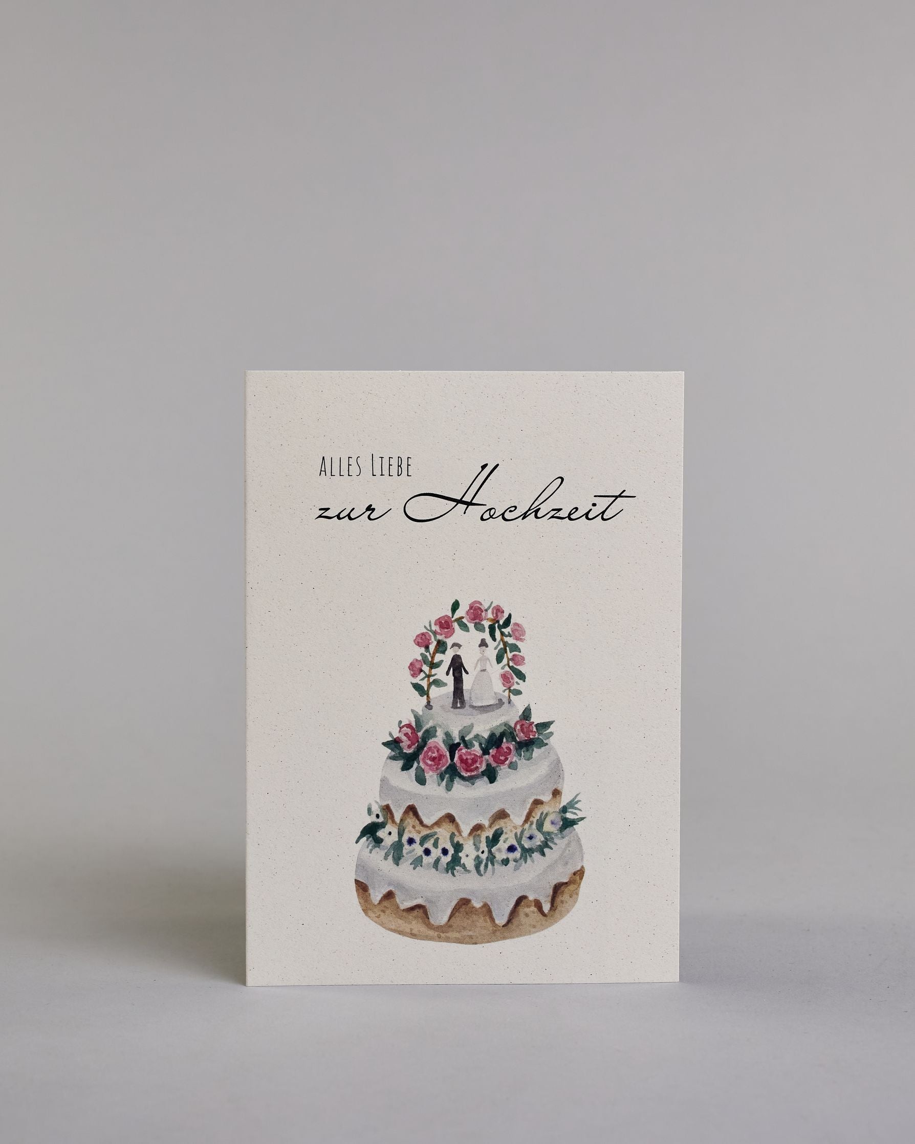 Glückwunschkarte zur Hochzeit mit schwarzer Kalligrafieaufschrift "Alles Liebe zur Hochzeit", darunter eine dreistückige Hochzeitstorte mit rosanen und weißen Blumen geschmückt und einem Brautpaar unter einem Hochzeitsbogen auf der obersten Tortenebene. Der Text "ALLES LIEBE" ist in dünner schwarzer Kalligraphieschrift, darunter das "zur Hochzeit" in geschwungener schwarzer Schreibschrift und alles ist auf hellem Hintergrund. Dazu ein weißes handgeschöpftes Kuvert. #1