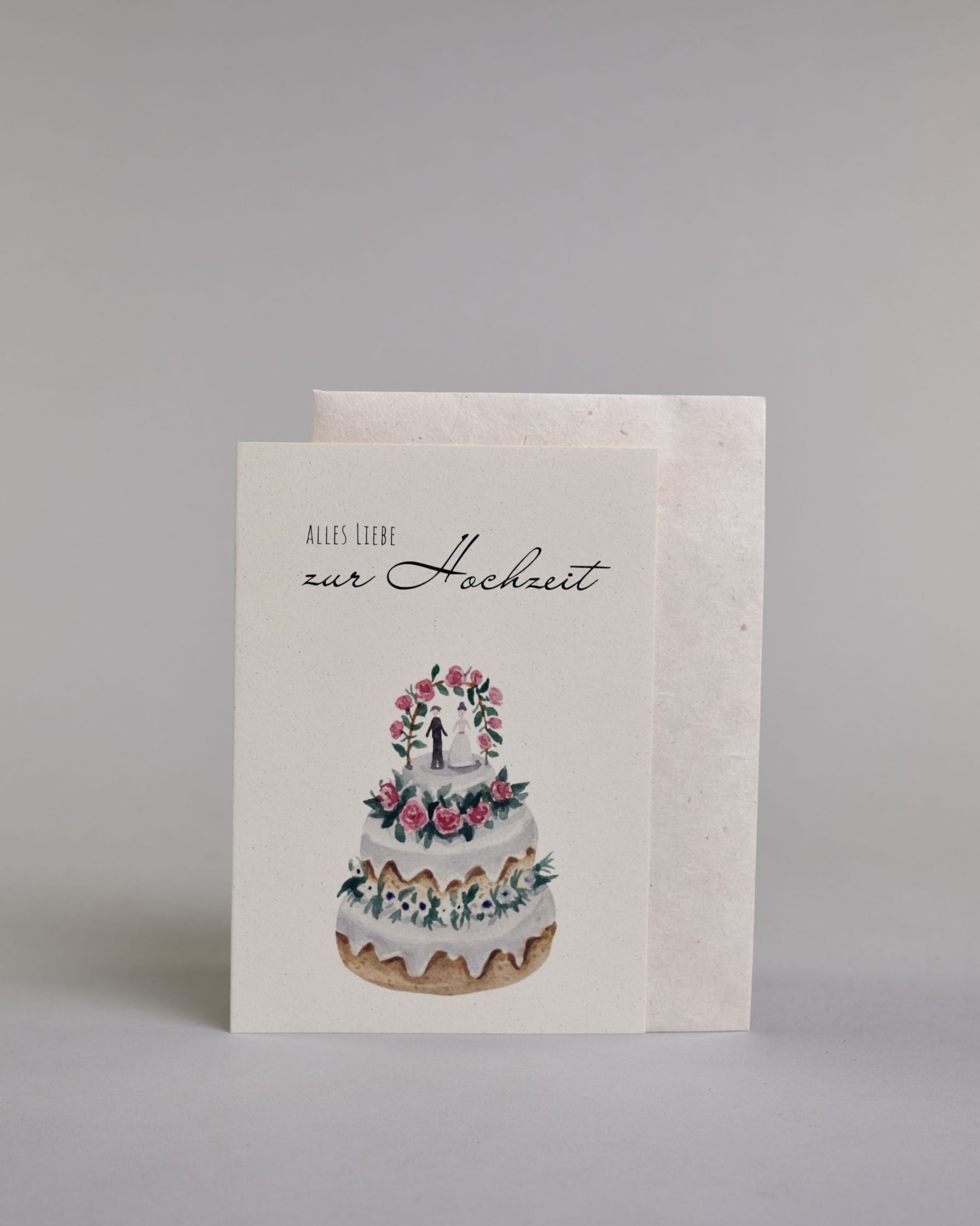 Glückwunschkarte zur Hochzeit mit schwarzer Kalligrafieaufschrift "Alles Liebe zur Hochzeit", darunter eine dreistückige Hochzeitstorte mit rosanen und weißen Blumen geschmückt und einem Brautpaar unter einem Hochzeitsbogen auf der obersten Tortenebene. Der Text "ALLES LIEBE" ist in dünner schwarzer Kalligraphieschrift, darunter das "zur Hochzeit" in geschwungener schwarzer Schreibschrift und alles ist auf hellem Hintergrund. Dazu ein weißes handgeschöpftes Kuvert. #2