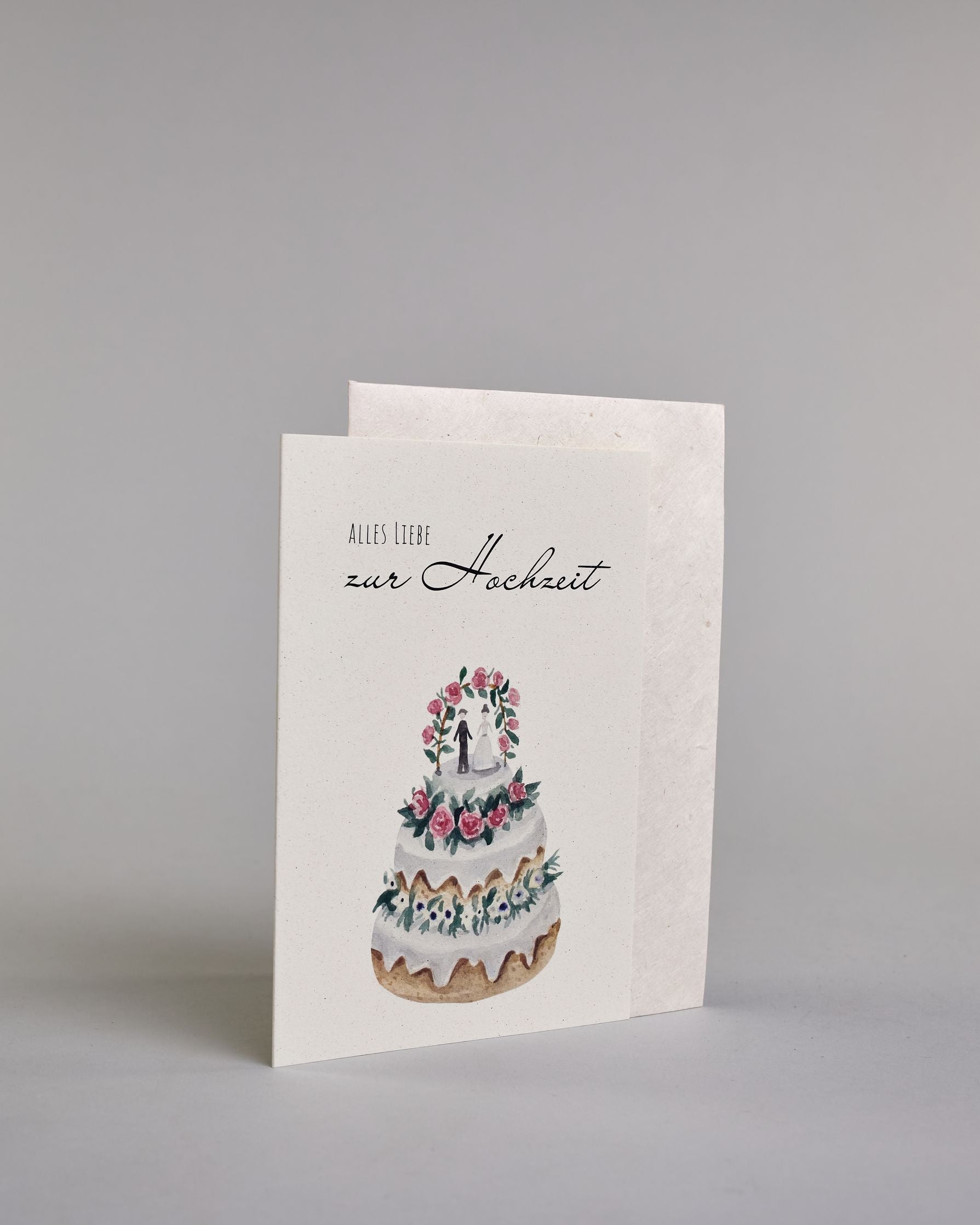 Glückwunschkarte zur Hochzeit mit schwarzer Kalligrafieaufschrift "Alles Liebe zur Hochzeit", darunter eine dreistückige Hochzeitstorte mit rosanen und weißen Blumen geschmückt und einem Brautpaar unter einem Hochzeitsbogen auf der obersten Tortenebene. Der Text "ALLES LIEBE" ist in dünner schwarzer Kalligraphieschrift, darunter das "zur Hochzeit" in geschwungener schwarzer Schreibschrift und alles ist auf hellem Hintergrund. Dazu ein weißes handgeschöpftes Kuvert. #3