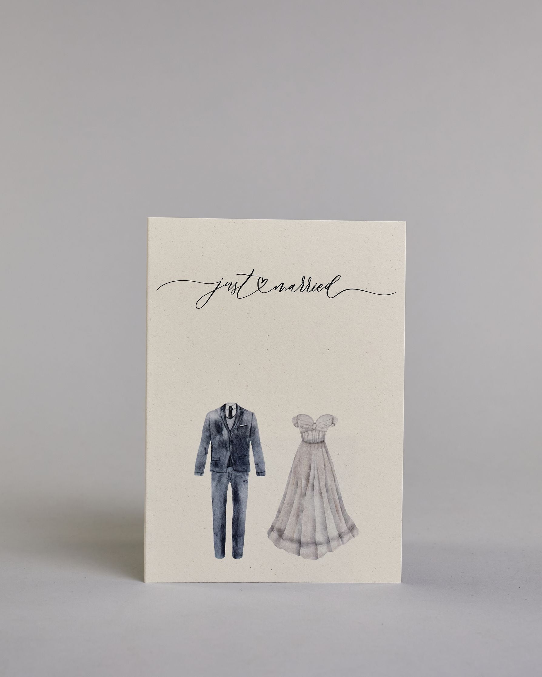 Glückwunschkarte auf Englisch zur Hochzeit mit schwarzer Kalligrafieaufschrift "just married", darunter ein schwarzer Anzug und rechts daneben ein weißes Hochzeitskleid. Der Text "just married" ist in geschwungener schwarzer Kalligraphieschrift und zieht sich von ganz links nach ganz rechts über die Karte, außerdem befindet sich im Schwung der Schrift in der Mitte der beiden Worte ein Herz und alles ist auf hellem Hintergrund. Dazu ein weißes handgeschöpftes Kuvert. #1